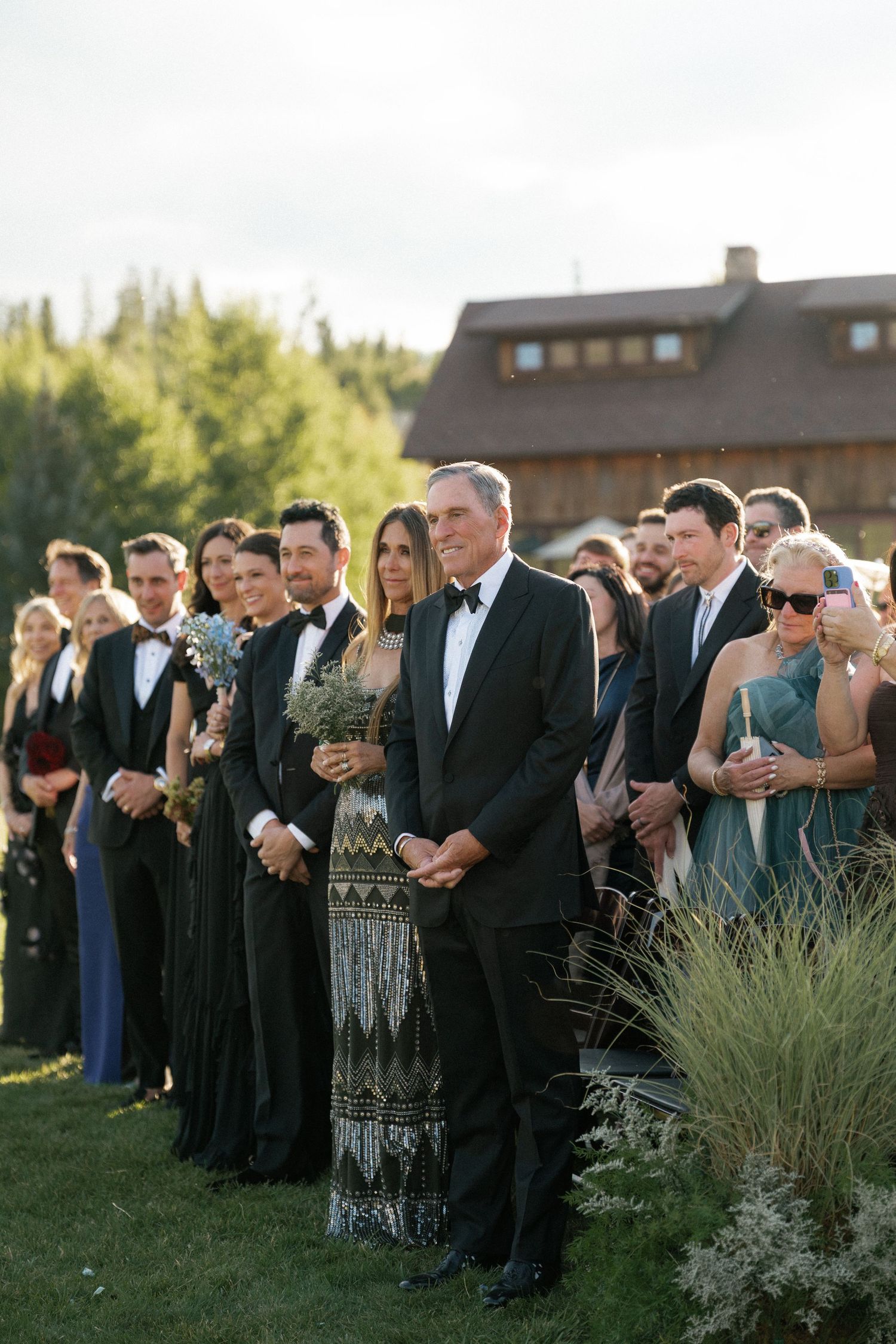 Wedding guests stand in formal attire during an outdoor ceremony.