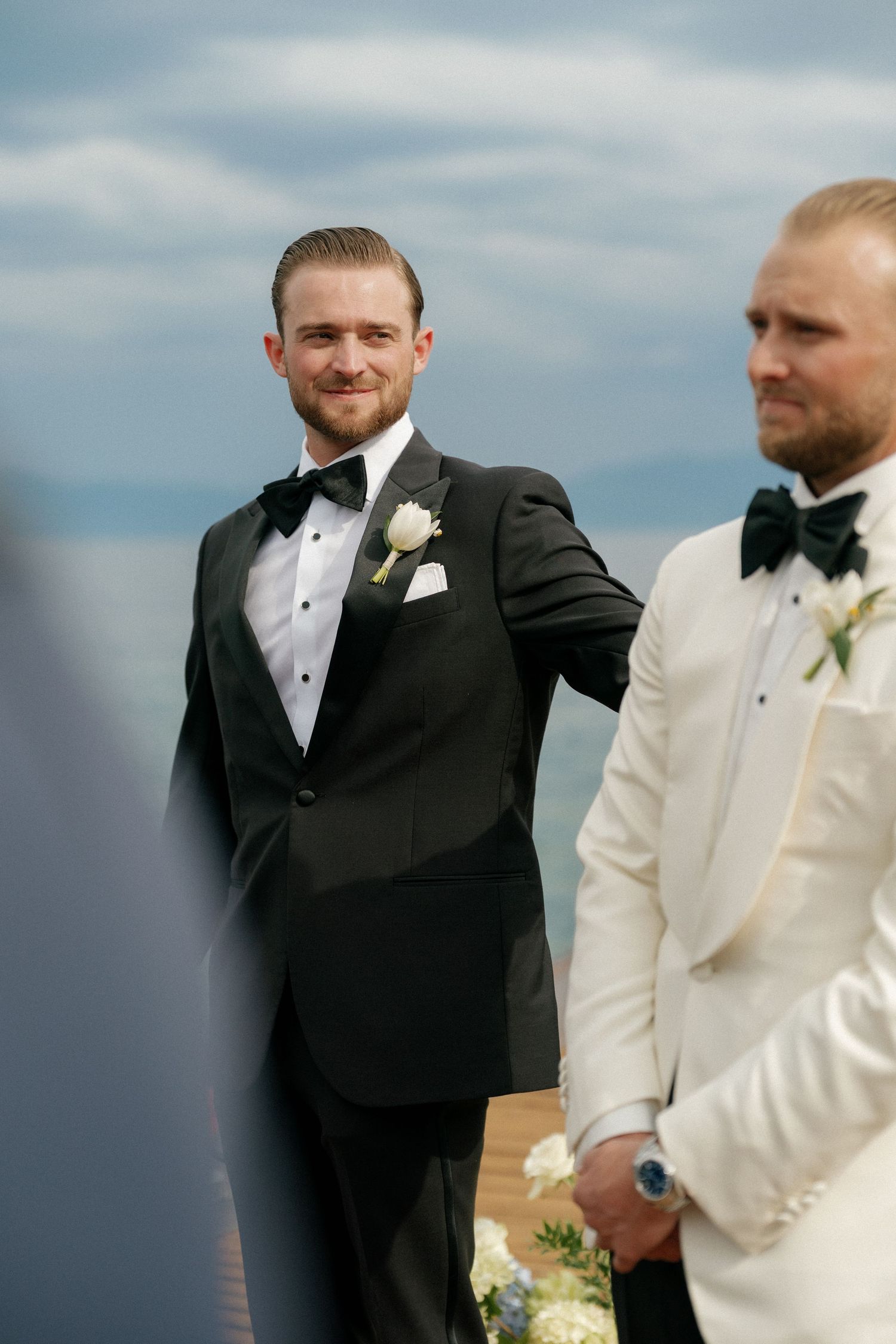 Groomsmen in black and white tuxedos standing at lakeside wedding ceremony.