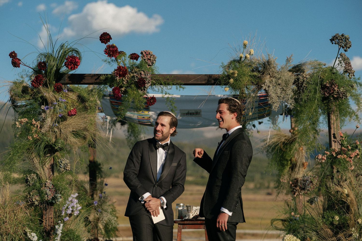 Wedding ceremony arch decorated with red flowers against a blue mountain sky.