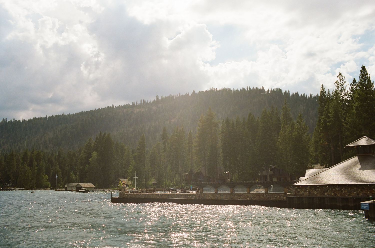 Lakeside mountain view with sunlight reflecting off the water and wooden buildings along the shore.