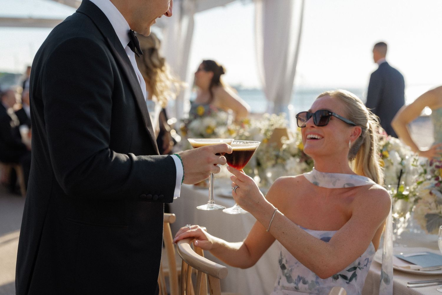 Smiling wedding guest in sunglasses toasting with red wine at an outdoor seaside reception.
