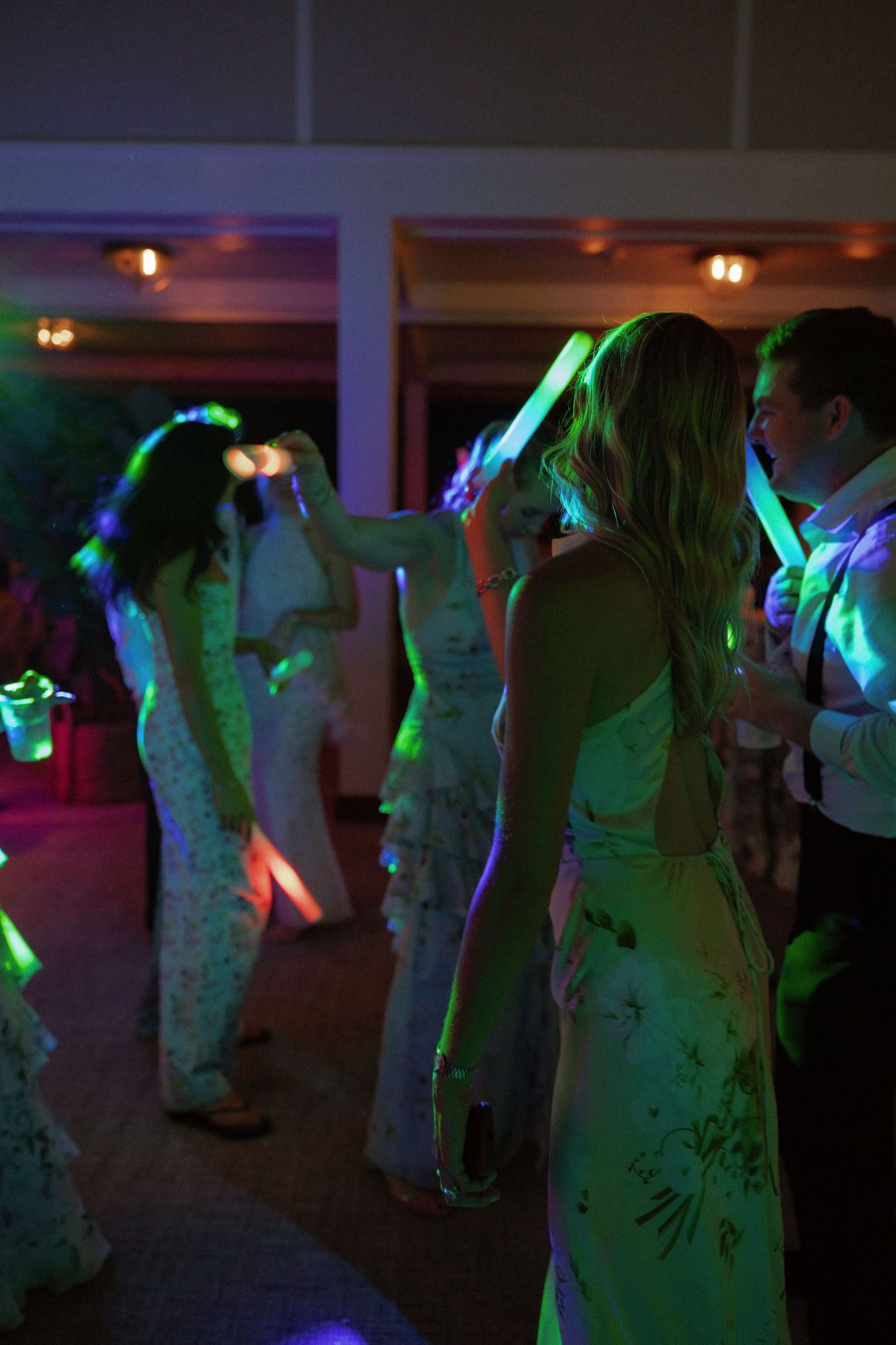 Guests dancing at a wedding reception with colorful neon glow sticks under vibrant green and purple party lights.