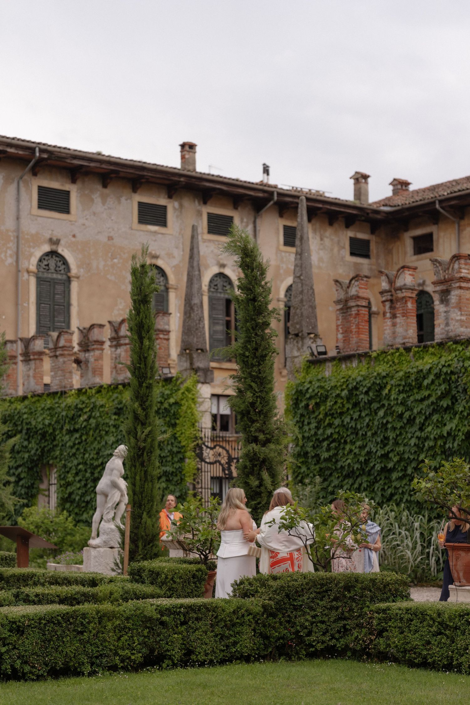 Guests explore a formal Italian garden with sculpted hedges and marble statues beside an ivy-covered historic villa.