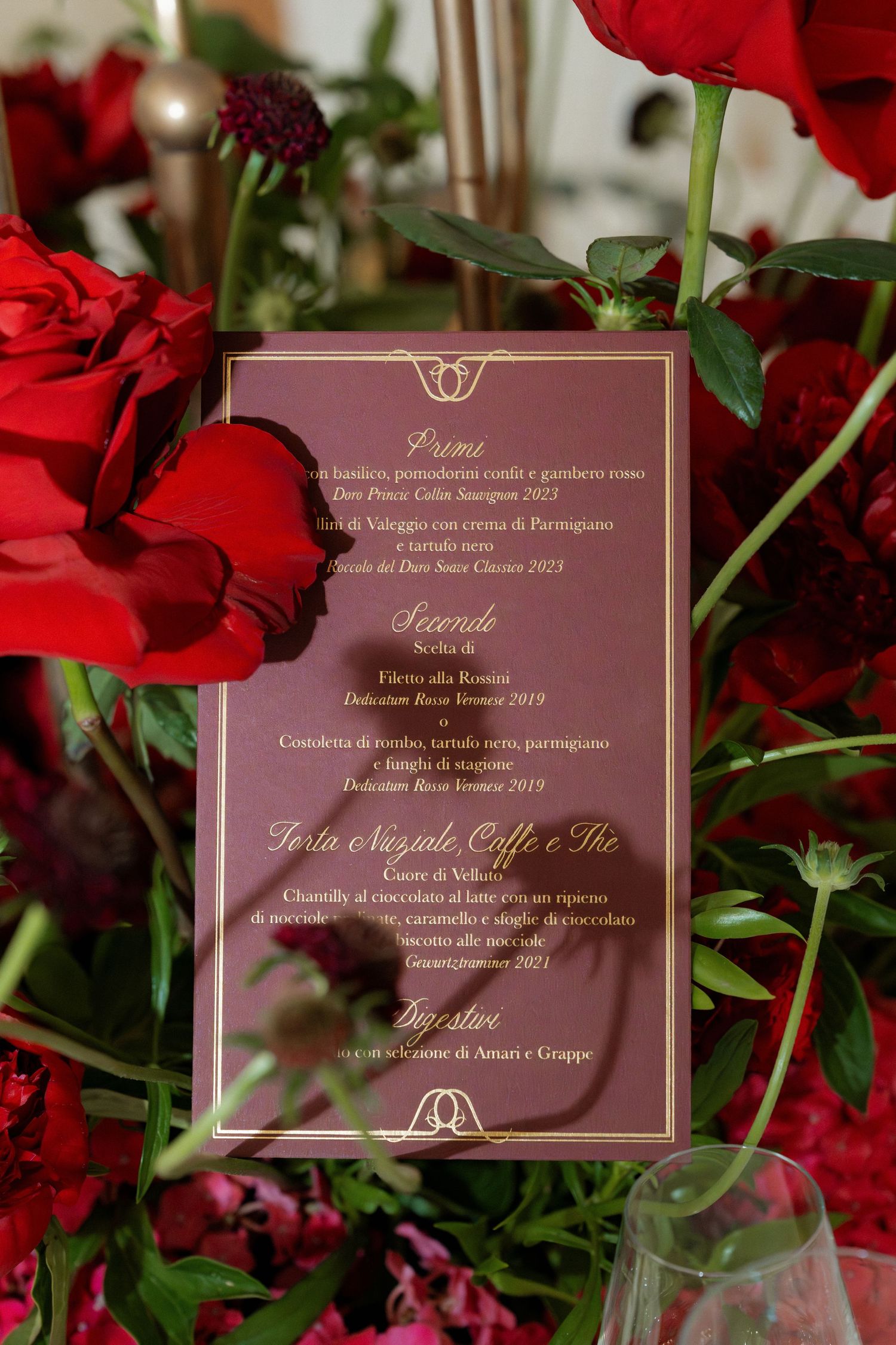 Elegant wedding menu card in burgundy and gold displayed among red roses and floral arrangements.