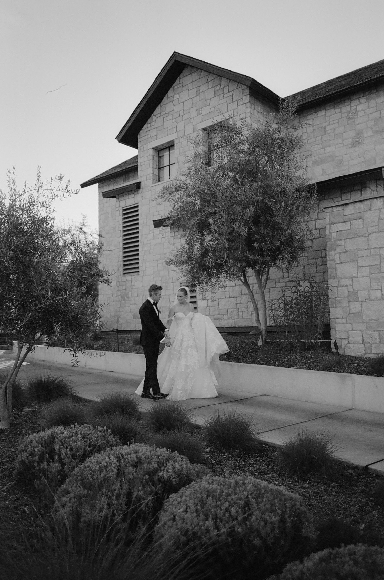 A couple poses in front of a modern stone building with landscaped gardens in a black and white photo.