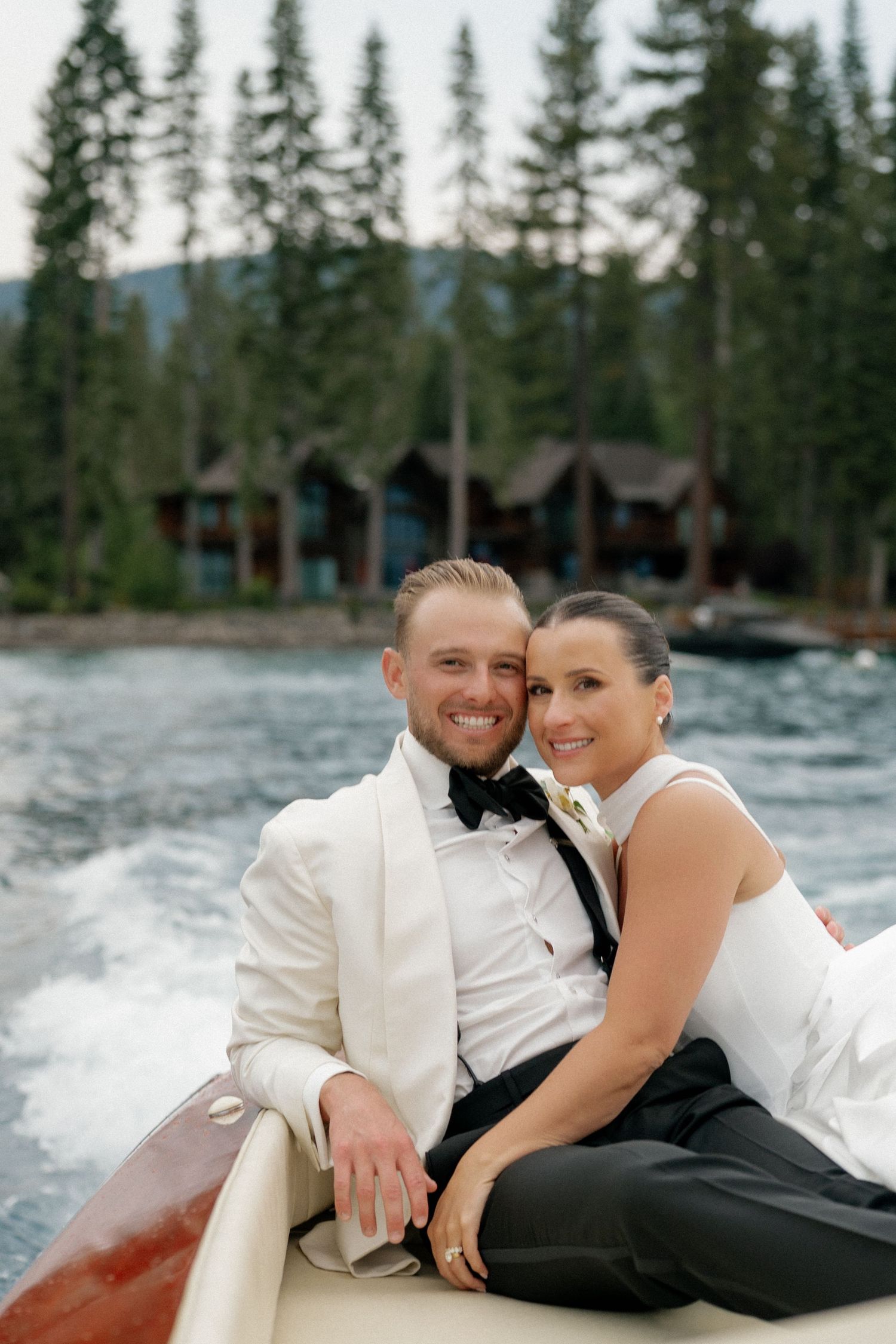 A romantic couple lounges together on a dock by a lake surrounded by evergreen trees.