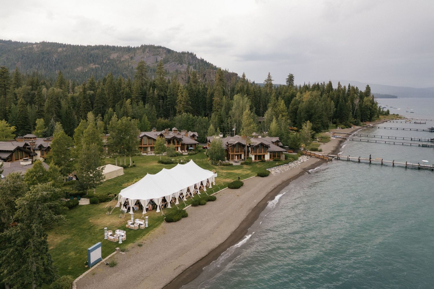 Aerial view of lakeside wedding venue with mountain backdrop and docked boats.