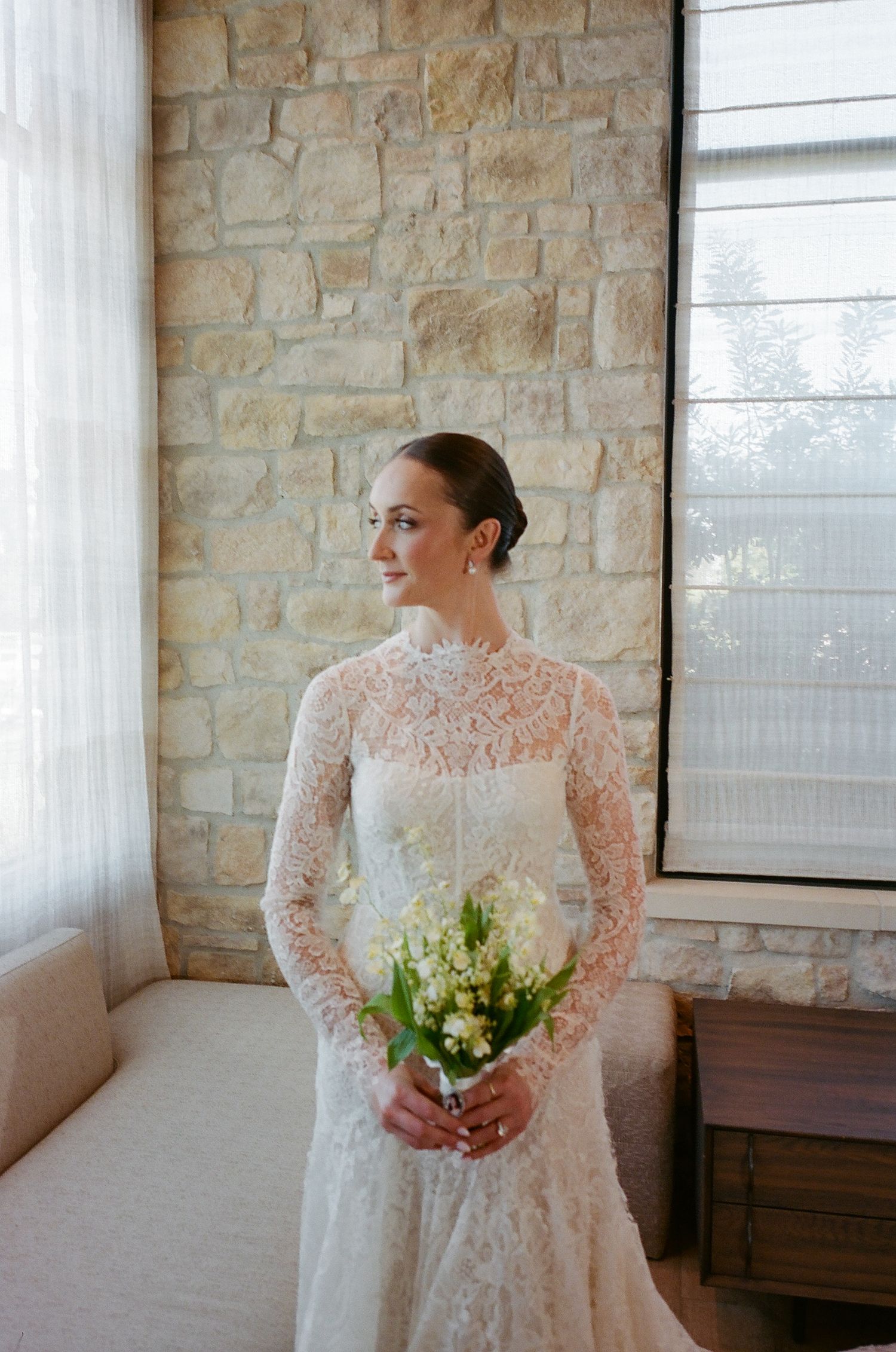 A bride in a delicate lace wedding gown stands in front of a stone wall holding a bouquet.