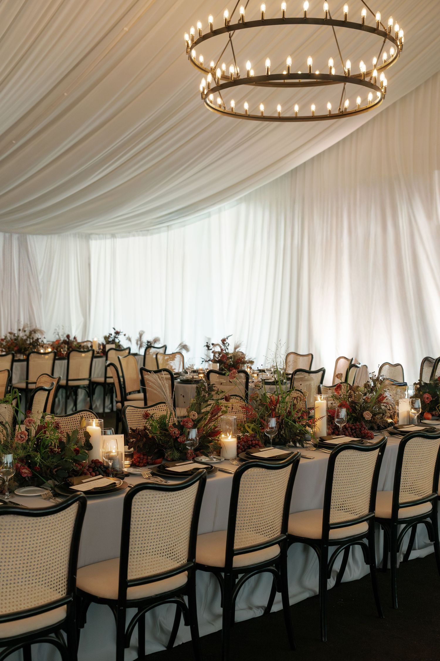 Elegant wedding reception tables set with cane back chairs and floral centerpieces under circular chandeliers in a draped tent.