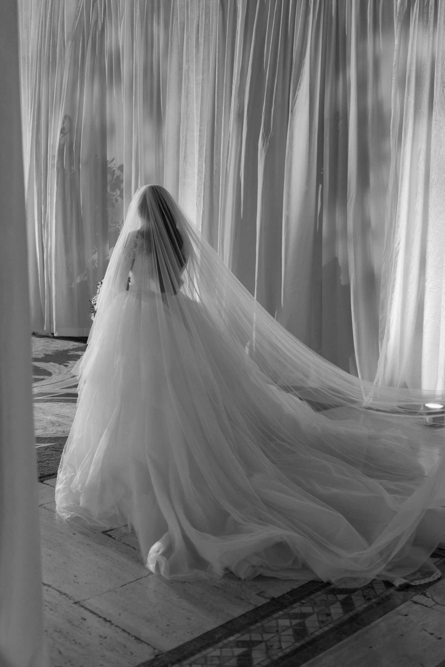 A long, flowing white wedding veil hangs in a room with curtains.