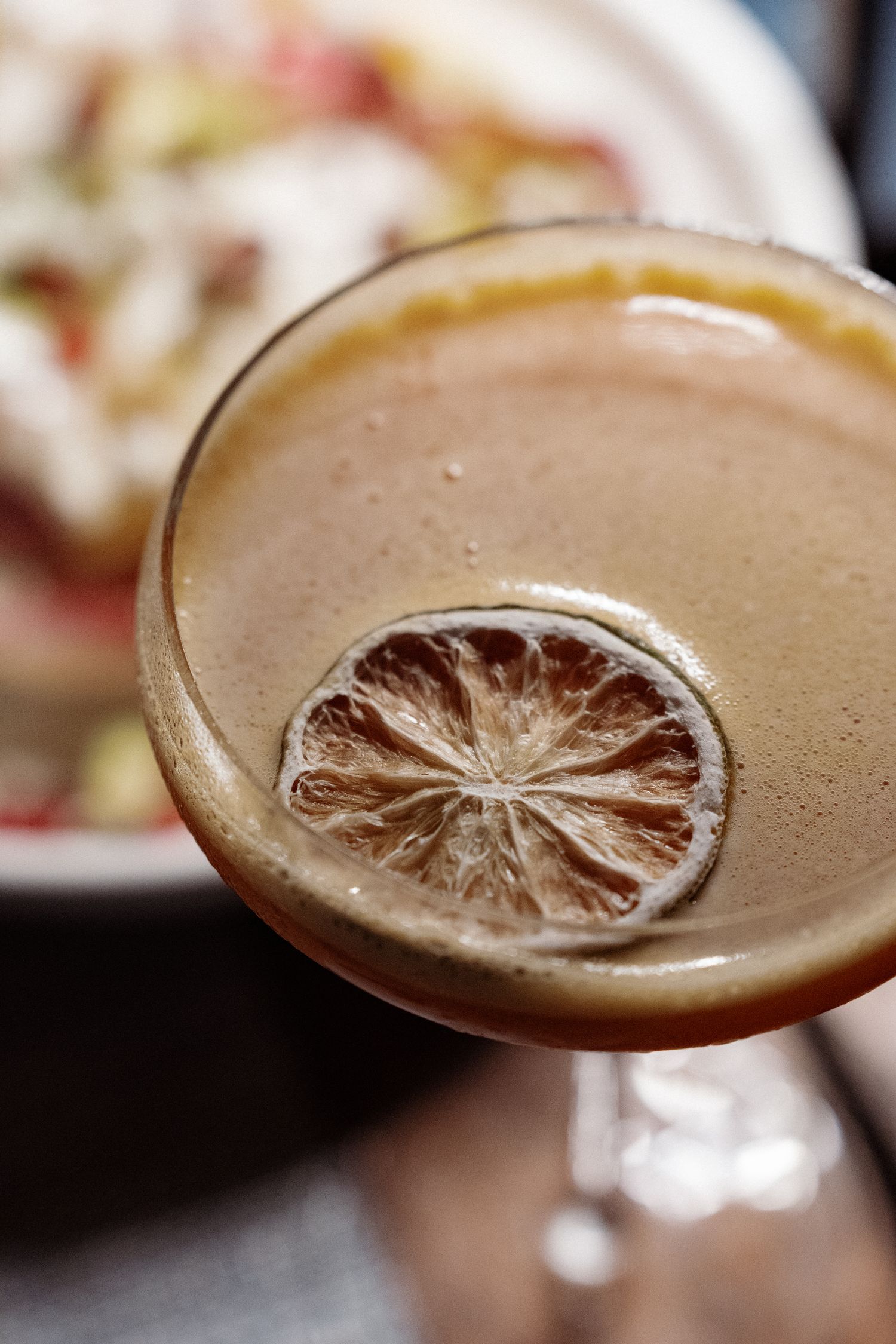 A close-up view of a glass containing a slice of lemon and a blurred background of food.
