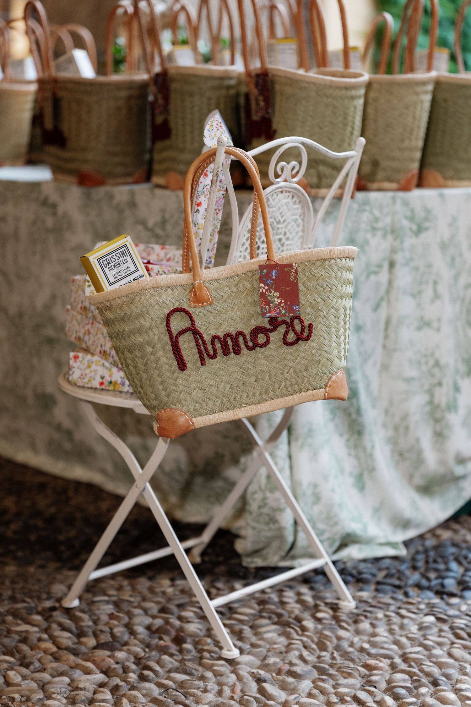 Personalized straw tote bag with leather handles, embroidered Amore text, displayed on white metal chair at wedding.