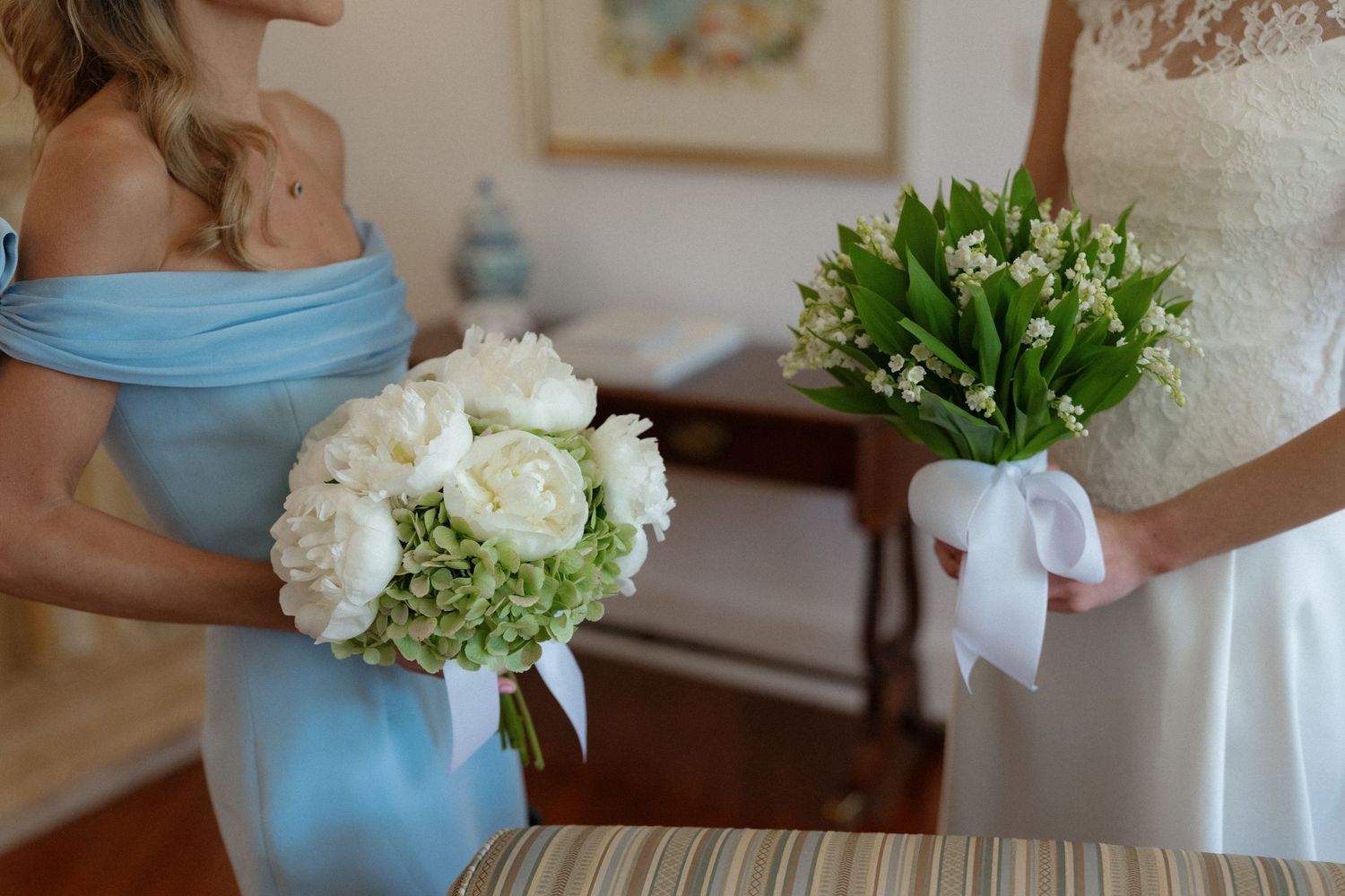 Bride and bridesmaid holding white floral bouquets, one with peonies and one with lily of the valley, at a wedding.