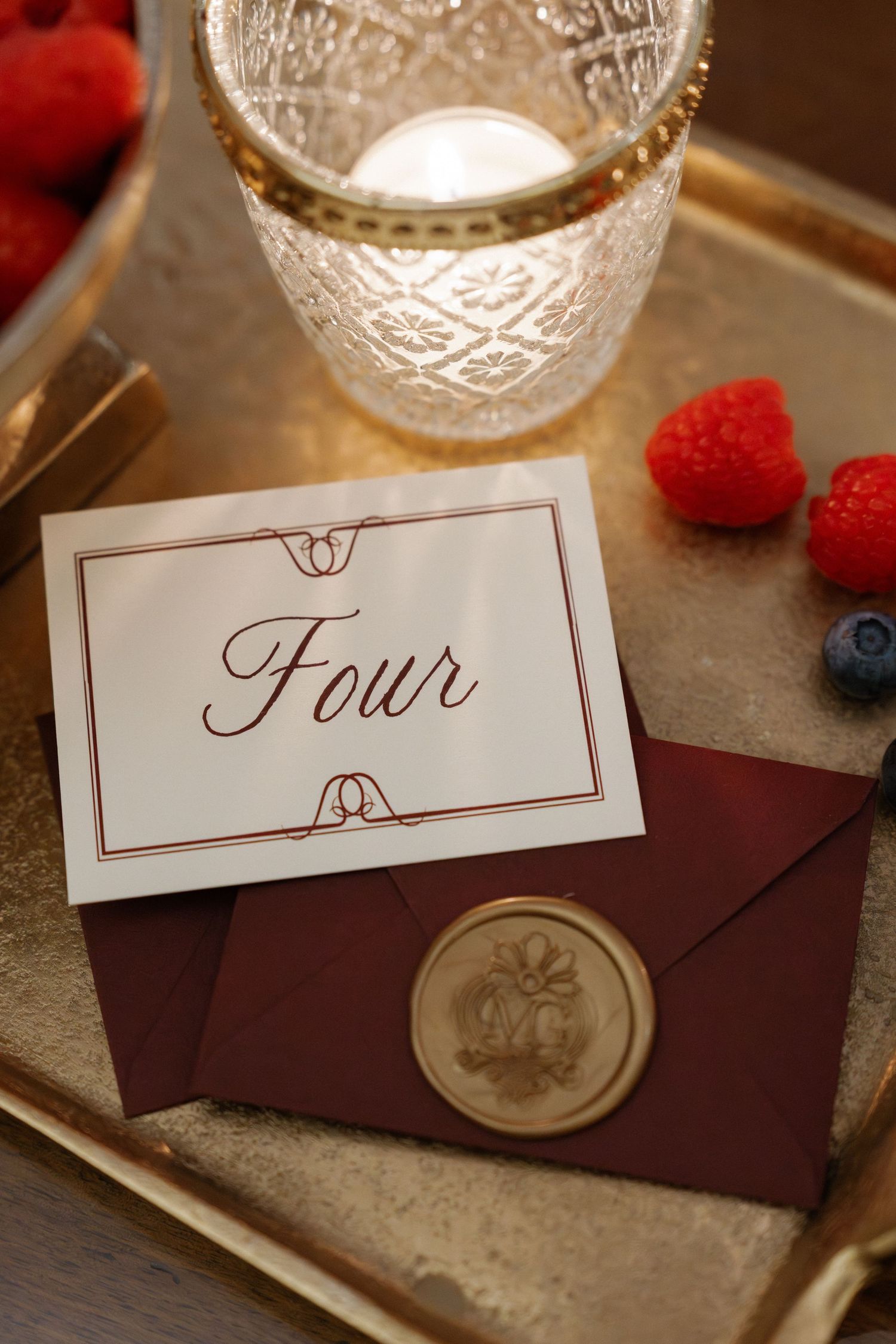 Wedding table number four card on burgundy envelope with wax seal, crystal candle holder and fresh berries on gold tray.