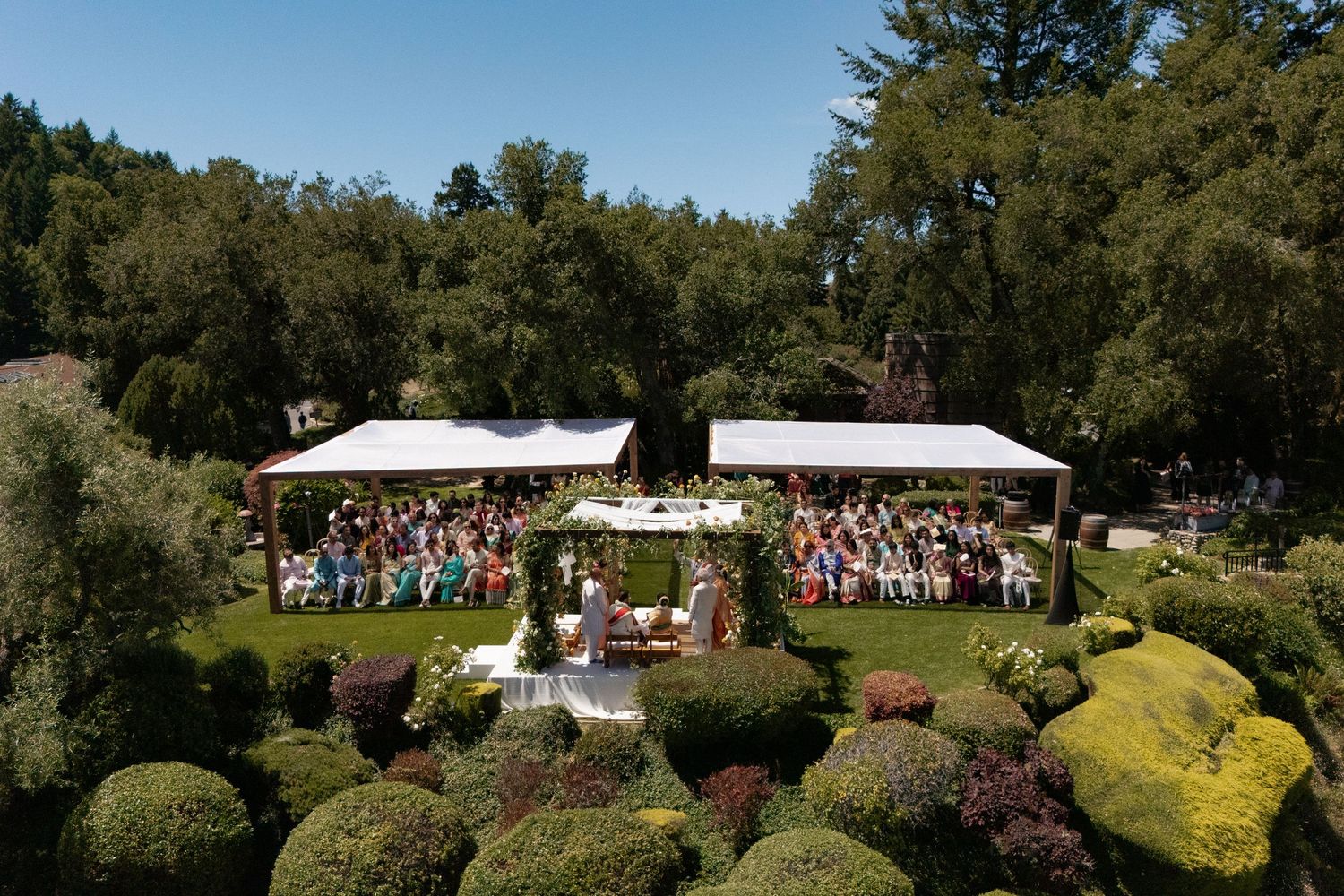 Aerial view of an elegant outdoor wedding ceremony with white canopies surrounded by lush green gardens and tall trees.