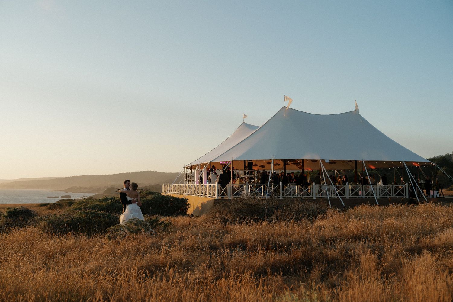 Large white wedding tent set up on coastal hillside during golden hour.
