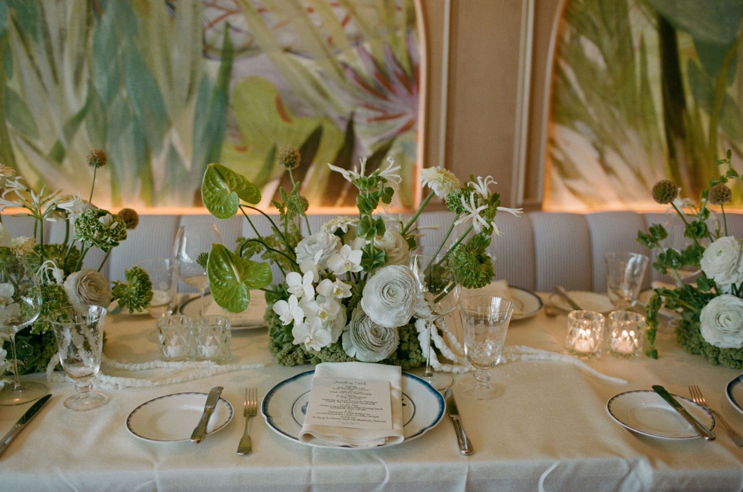 Elegant table setting with white roses and greenery centerpieces for wedding reception.