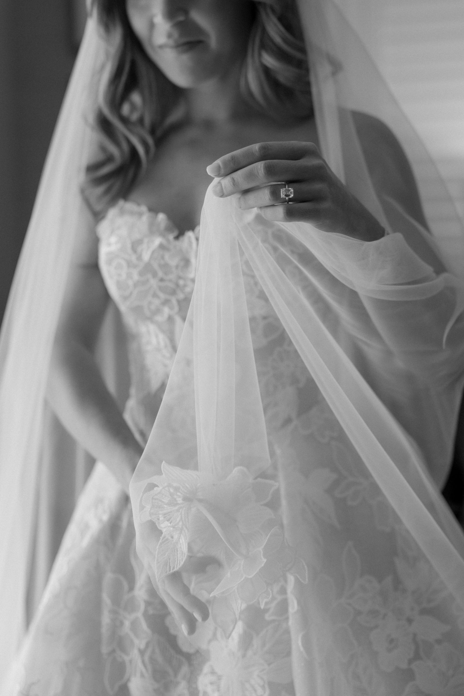 Close-up black and white detail of a lace wedding dress and veil with an engagement ring.