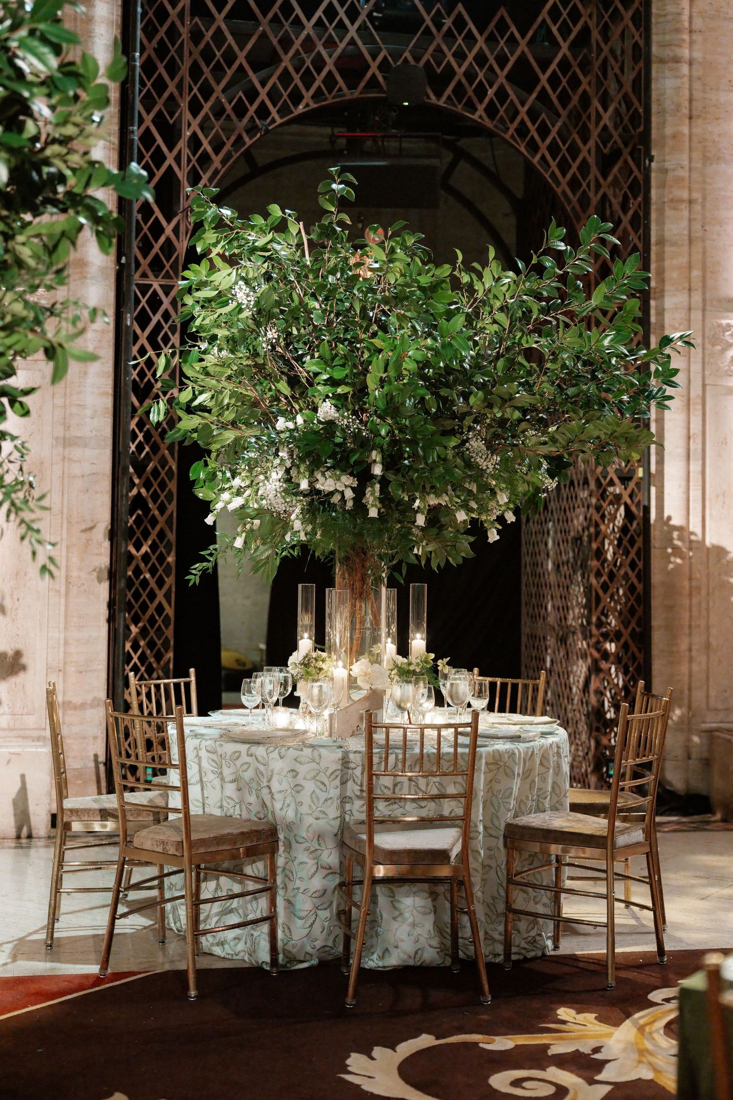 A table set for a formal event, with a floral centerpiece and candles, in a room with arched windows and greenery.