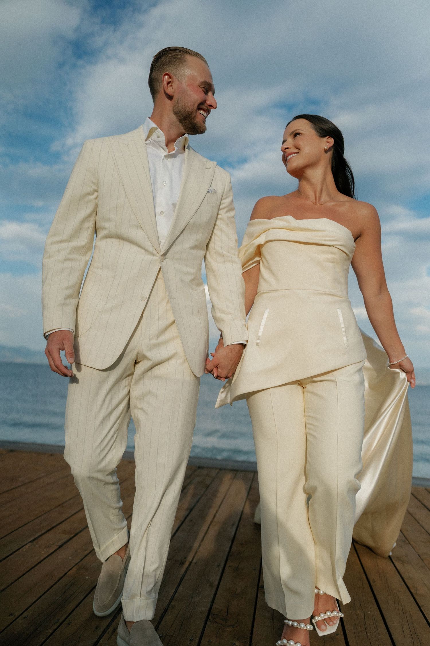 Couple in matching cream-colored outfits walking together on a wooden dock by the water.
