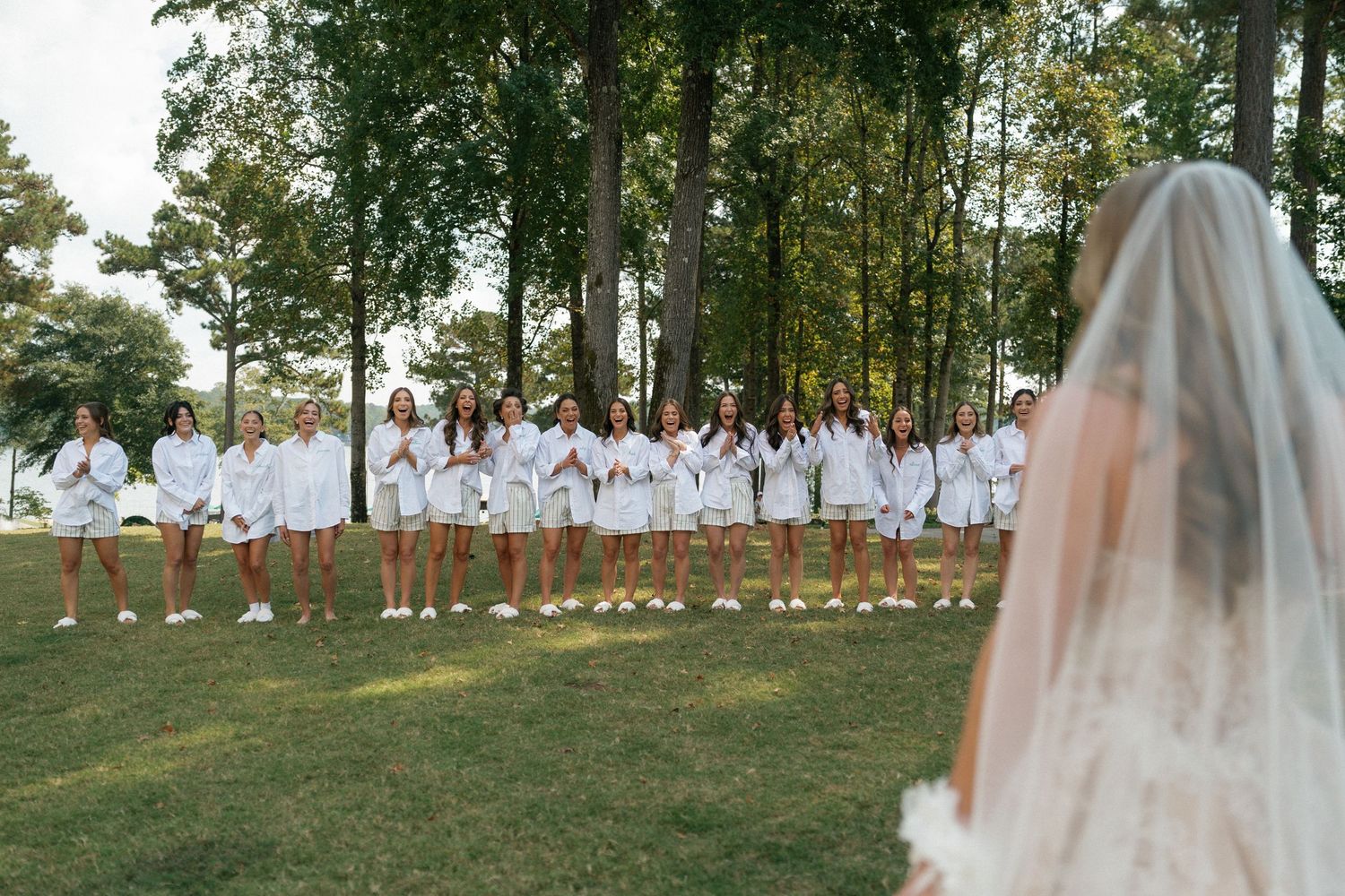 A wedding party in white dresses stands in a line on a grassy lawn as the bride looks on.