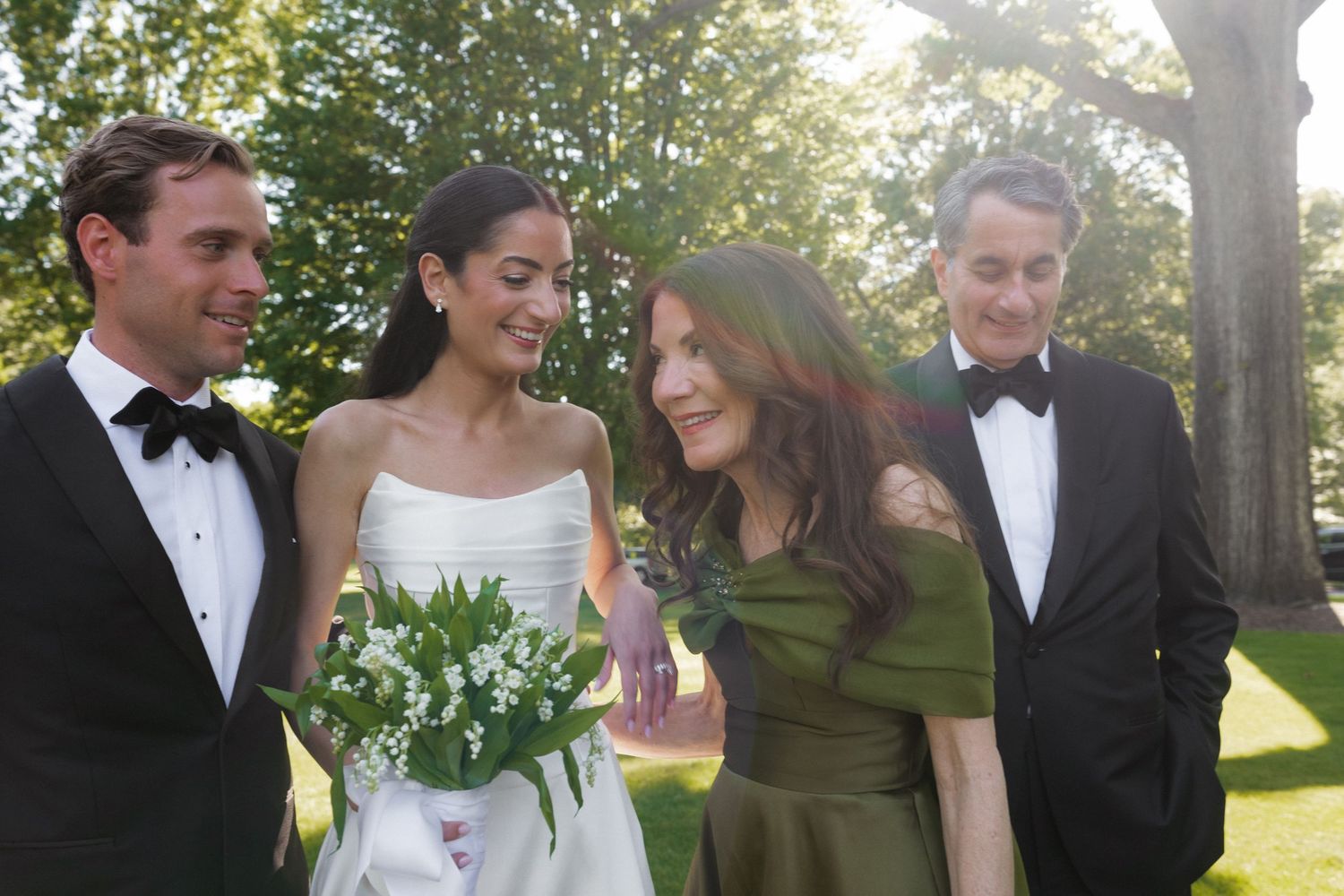 Bride and groom in black tuxedo pose with smiling family members in green dresses outdoors at wedding.