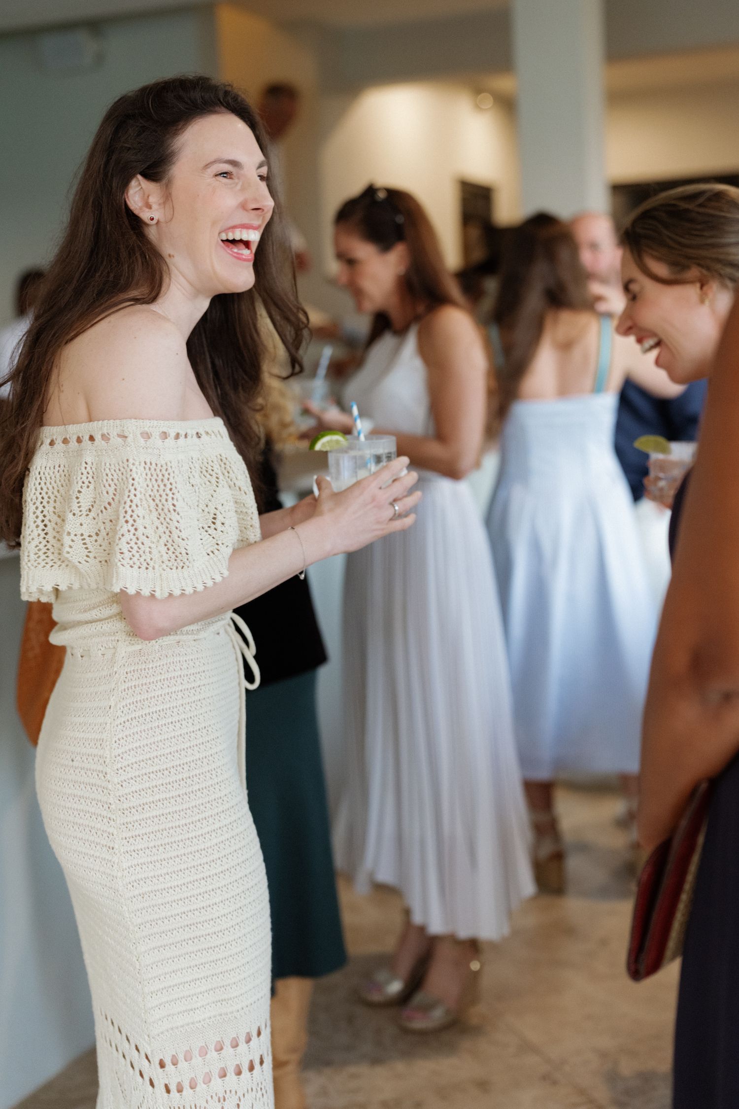 A woman in a white dress smiles and interacts with others at what appears to be a social event.
