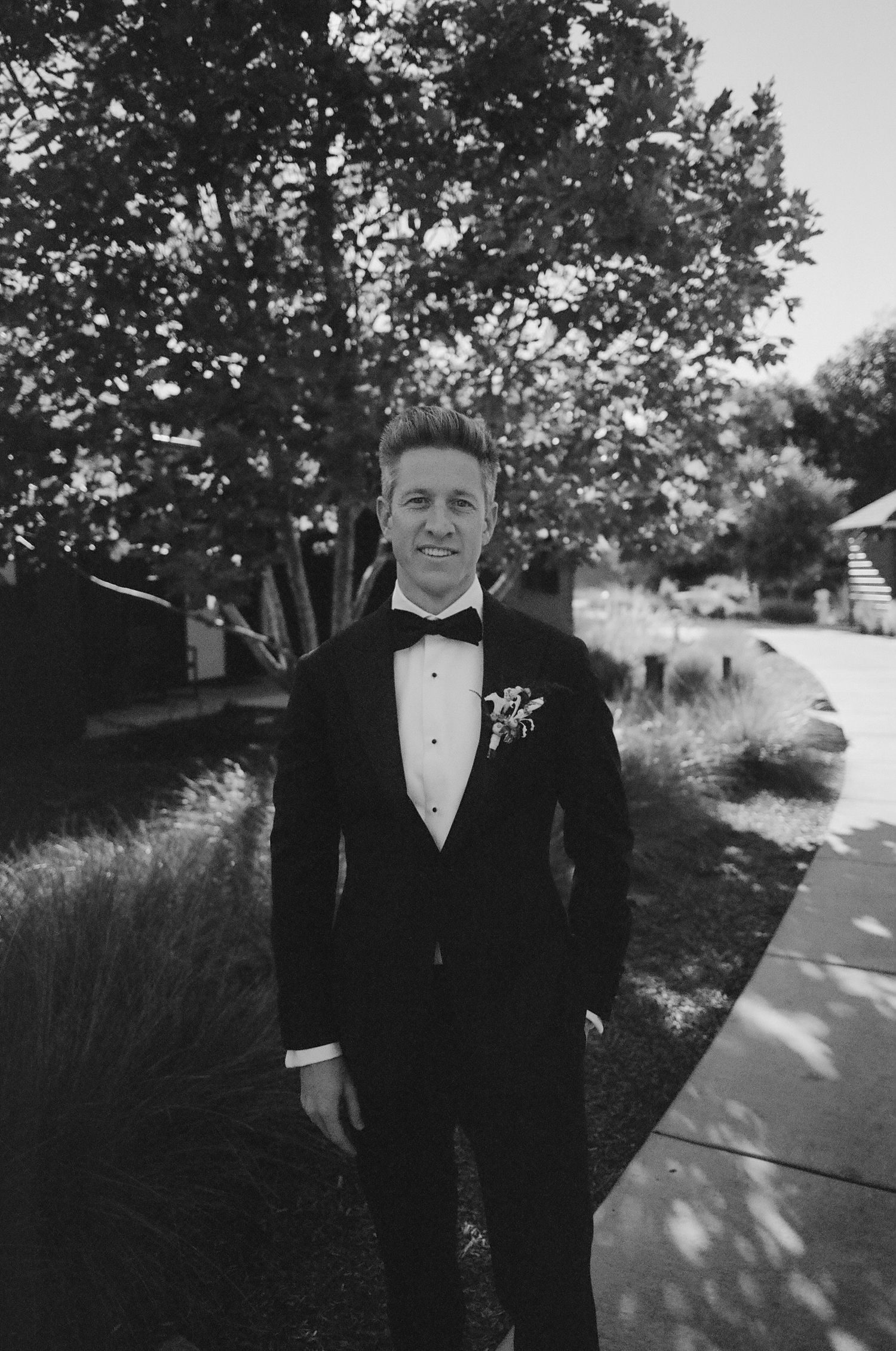 A groom in a classic black tuxedo poses outdoors near trees.