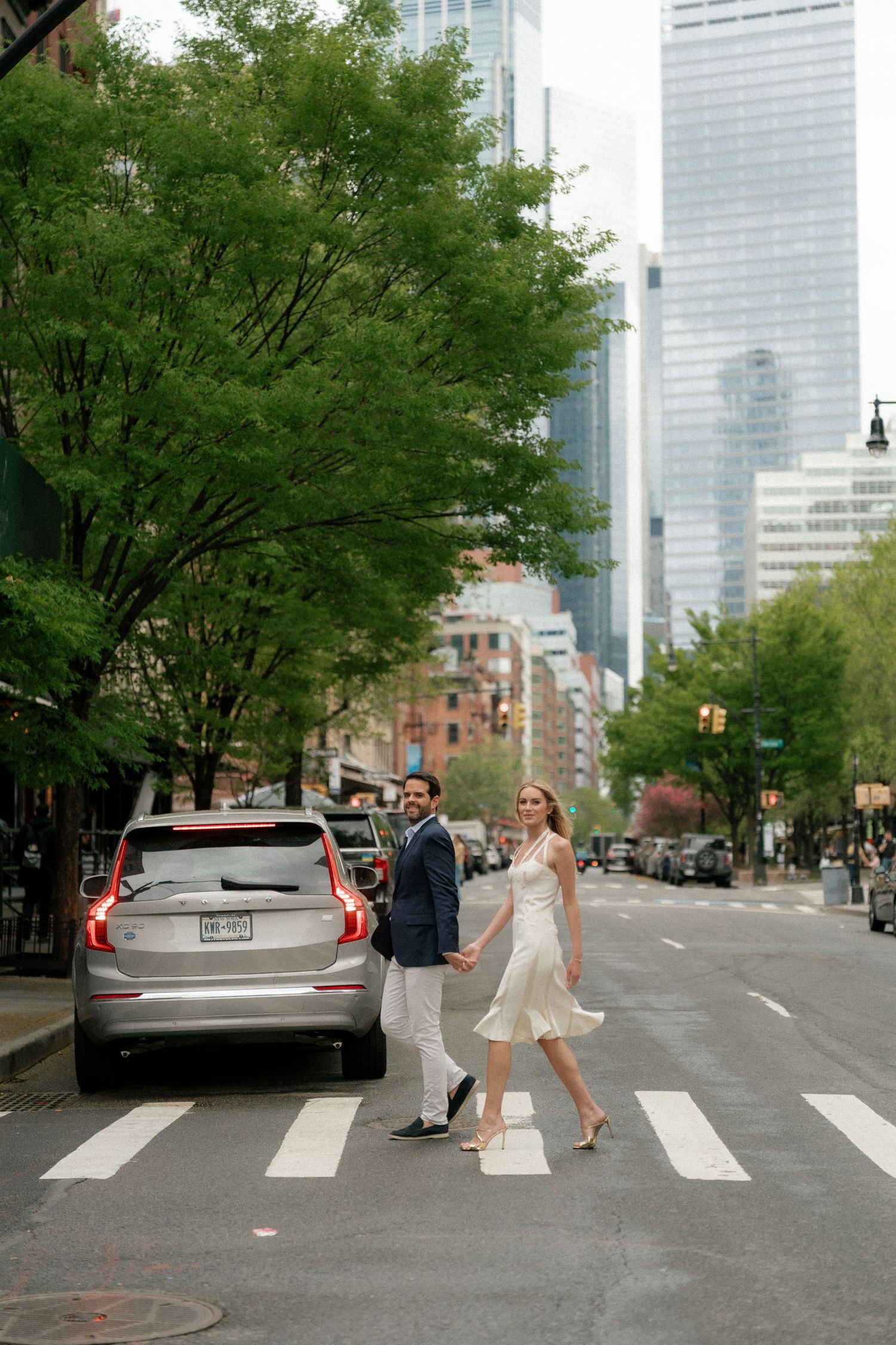 A couple walks across a city crosswalk with tall buildings and trees in the background.