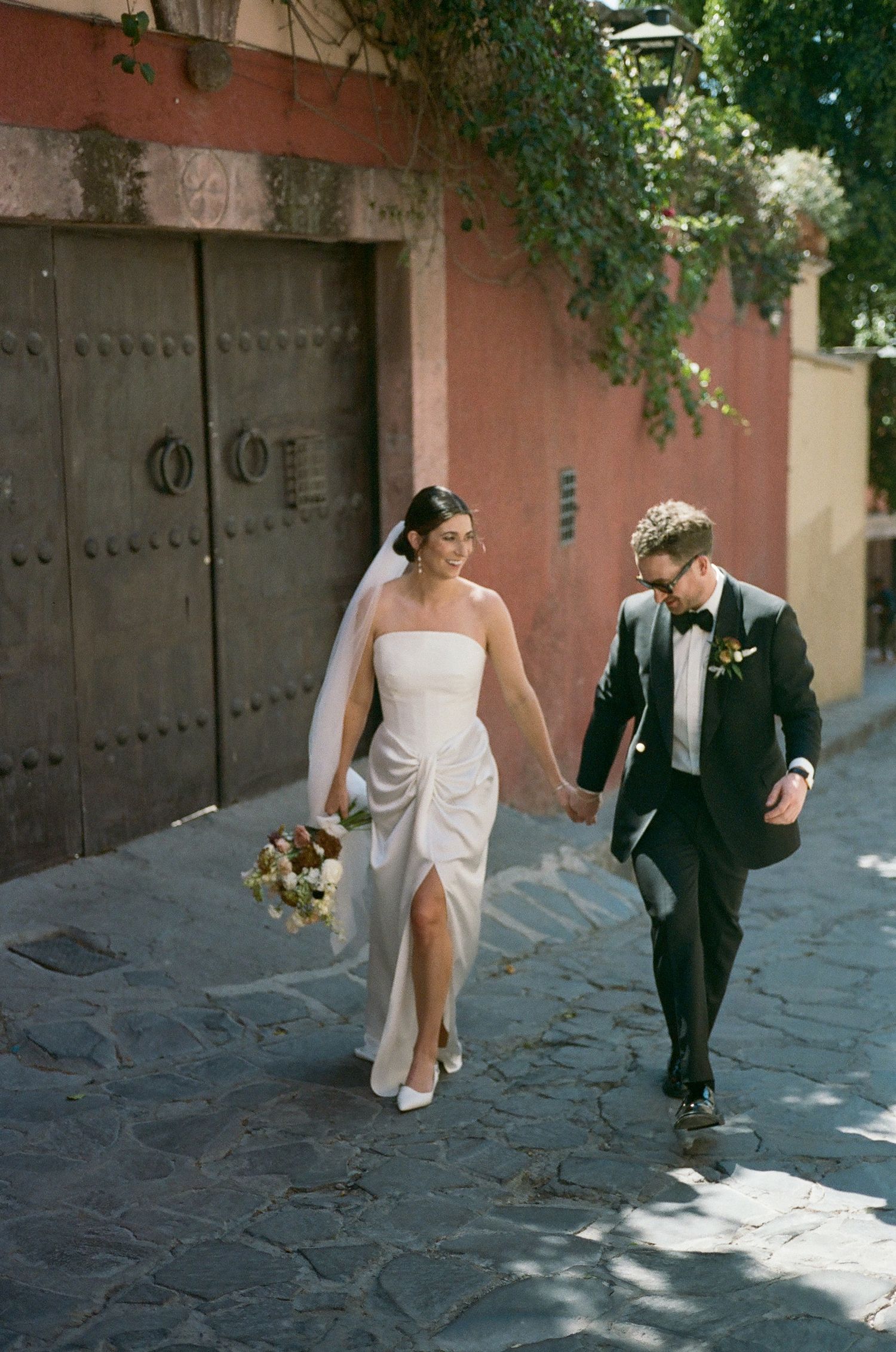Couple walking along a cobblestone street with pink walls and ornate wooden doors.