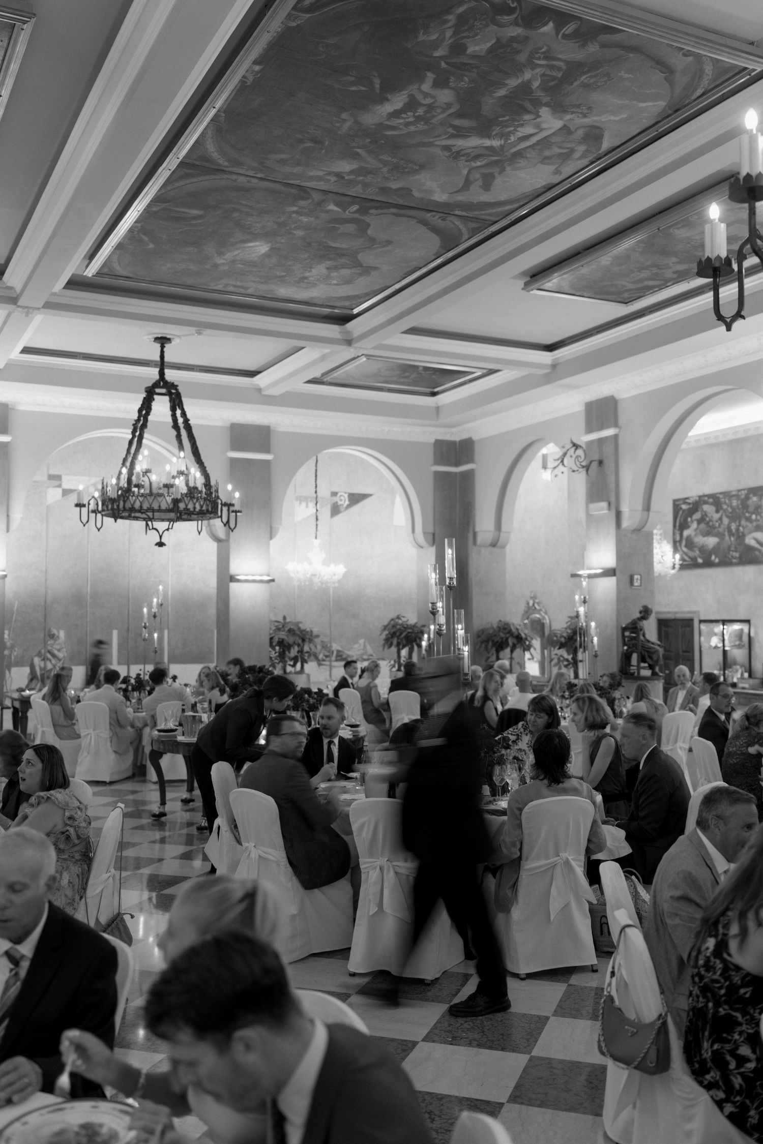 Black and white photo of guests dining in grand ballroom with ornate painted ceiling and chandelier.
