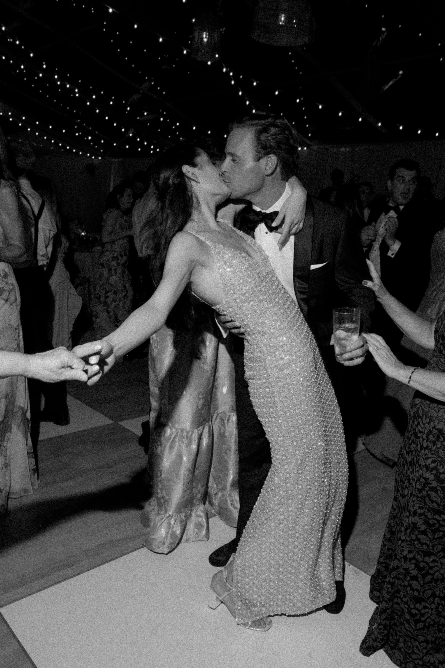 Couple sharing a kiss on the dance floor at a formal event, black and white photo with string lights above.