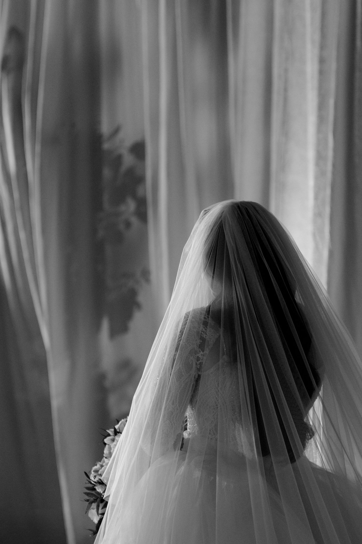 A black and white image of a bride wearing a long, flowing veil standing in front of a window.