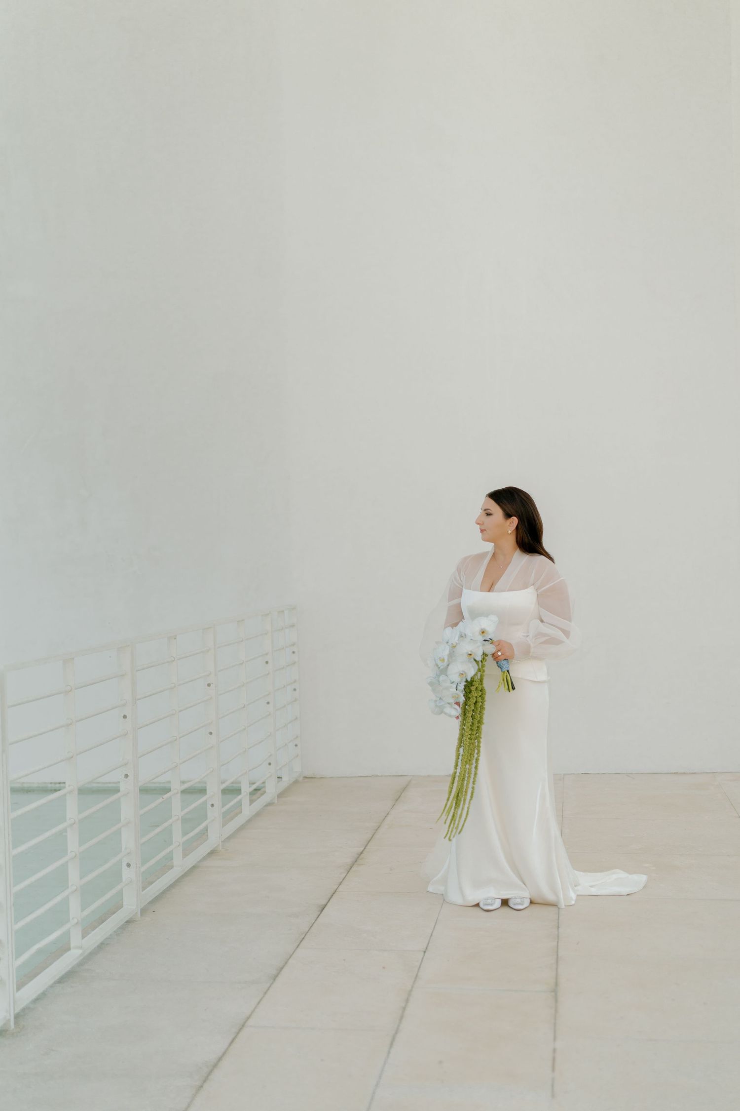 A bride in a flowing white dress and sheer sleeves stands elegantly against a minimalist white wall holding a bouquet.