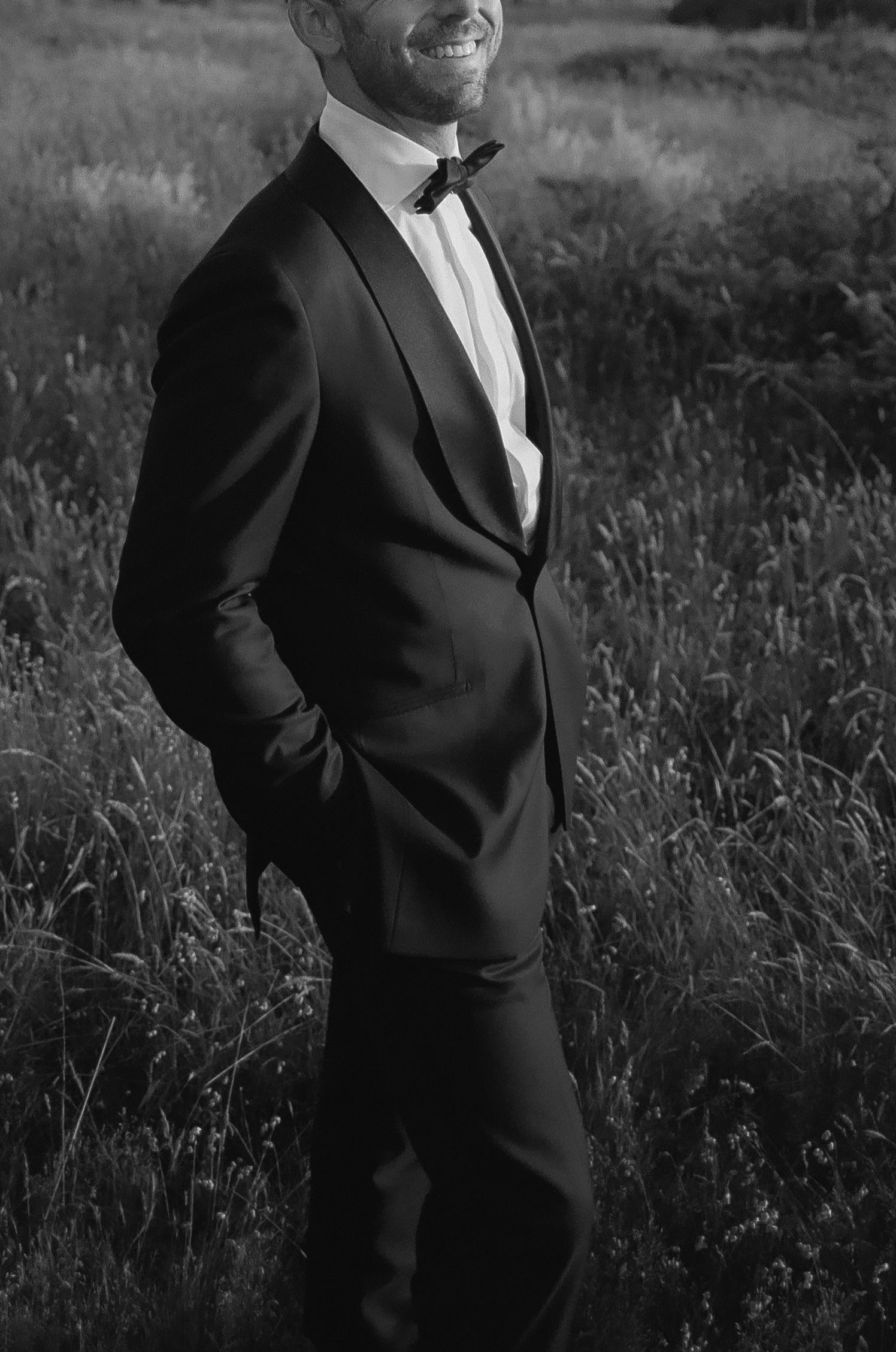 Person in black tuxedo standing in tall grass during an outdoor photoshoot.