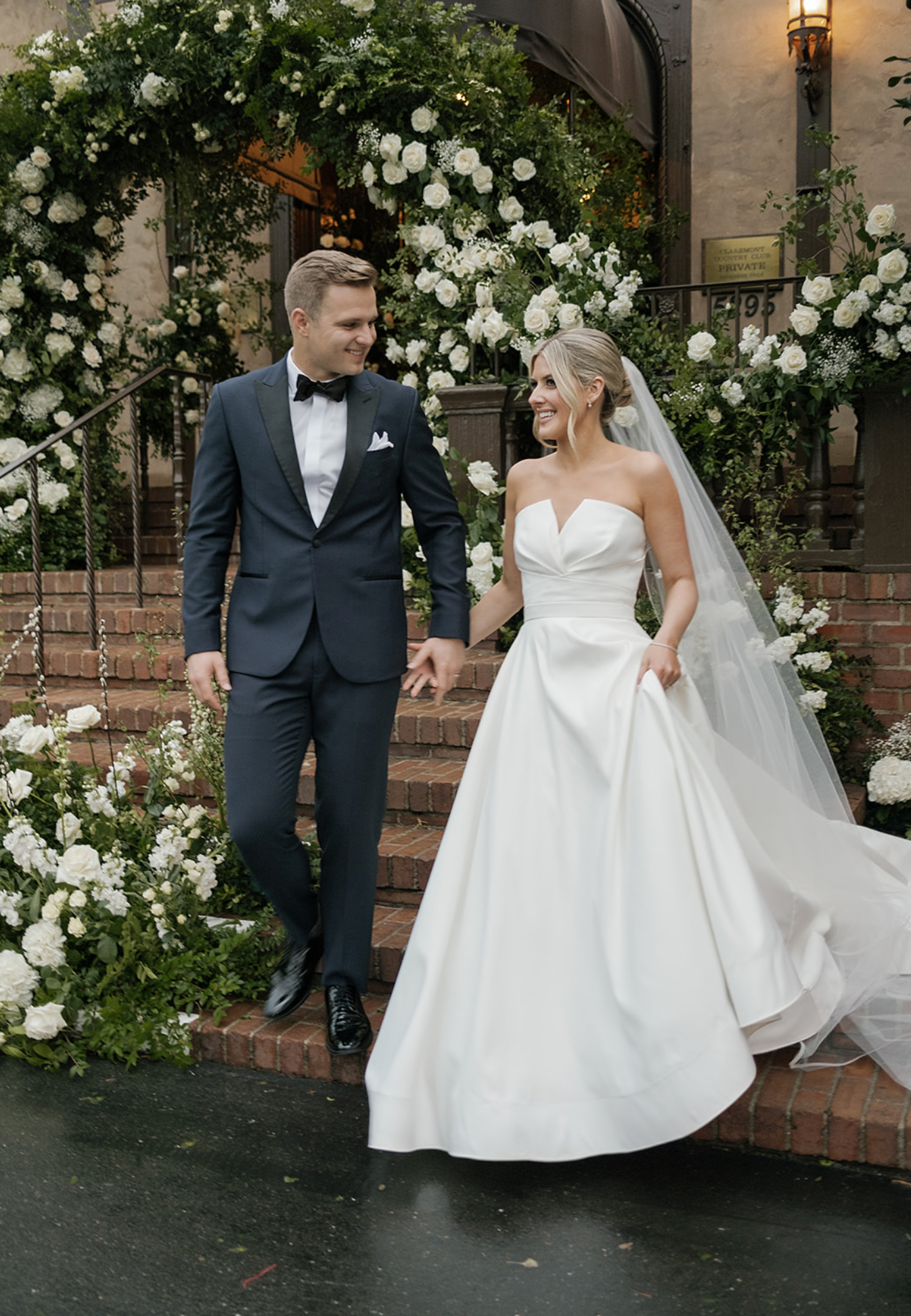 Wedding couple posing against a wall decorated with white roses and greenery.