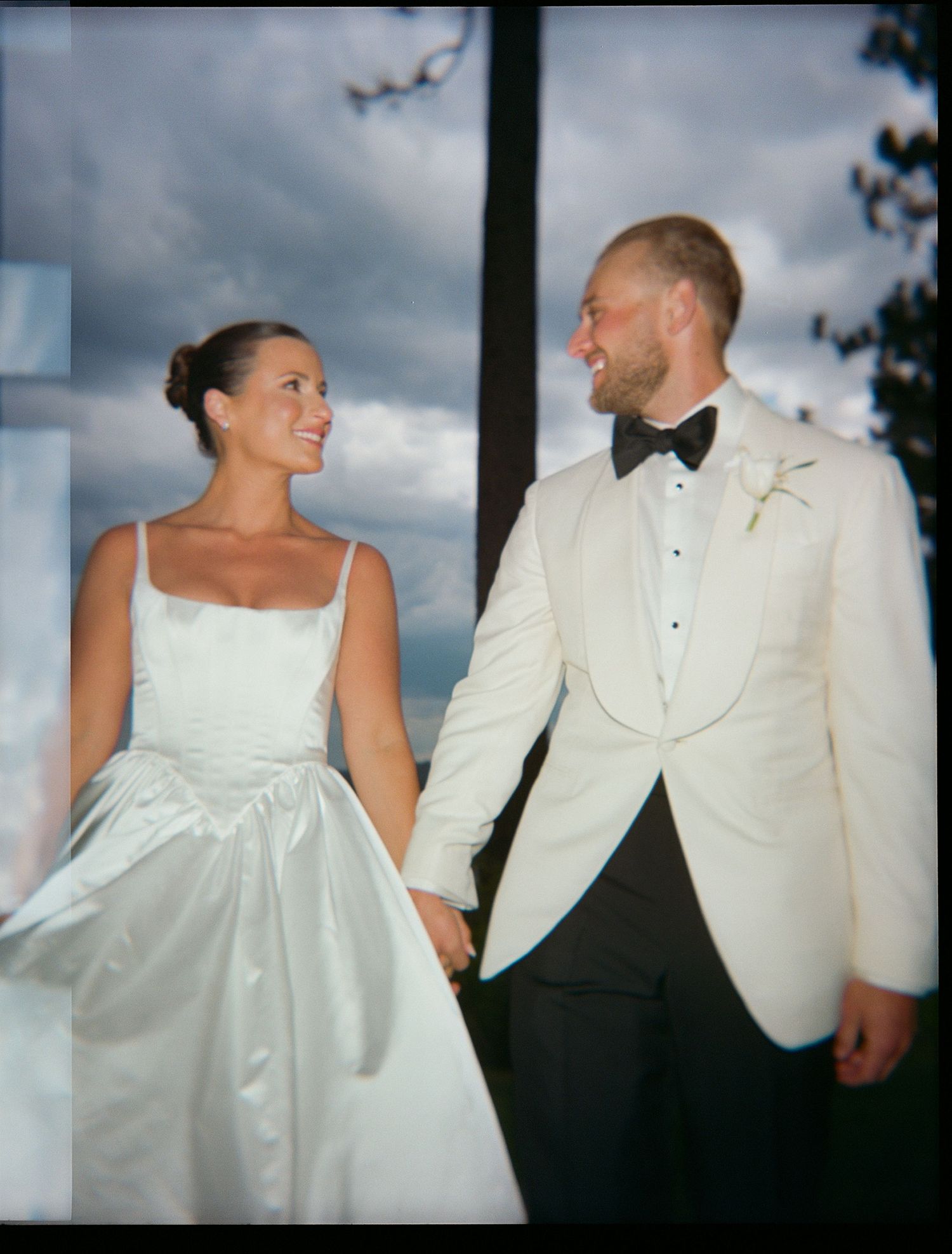 Couple in elegant white wedding attire standing together against cloudy sky.