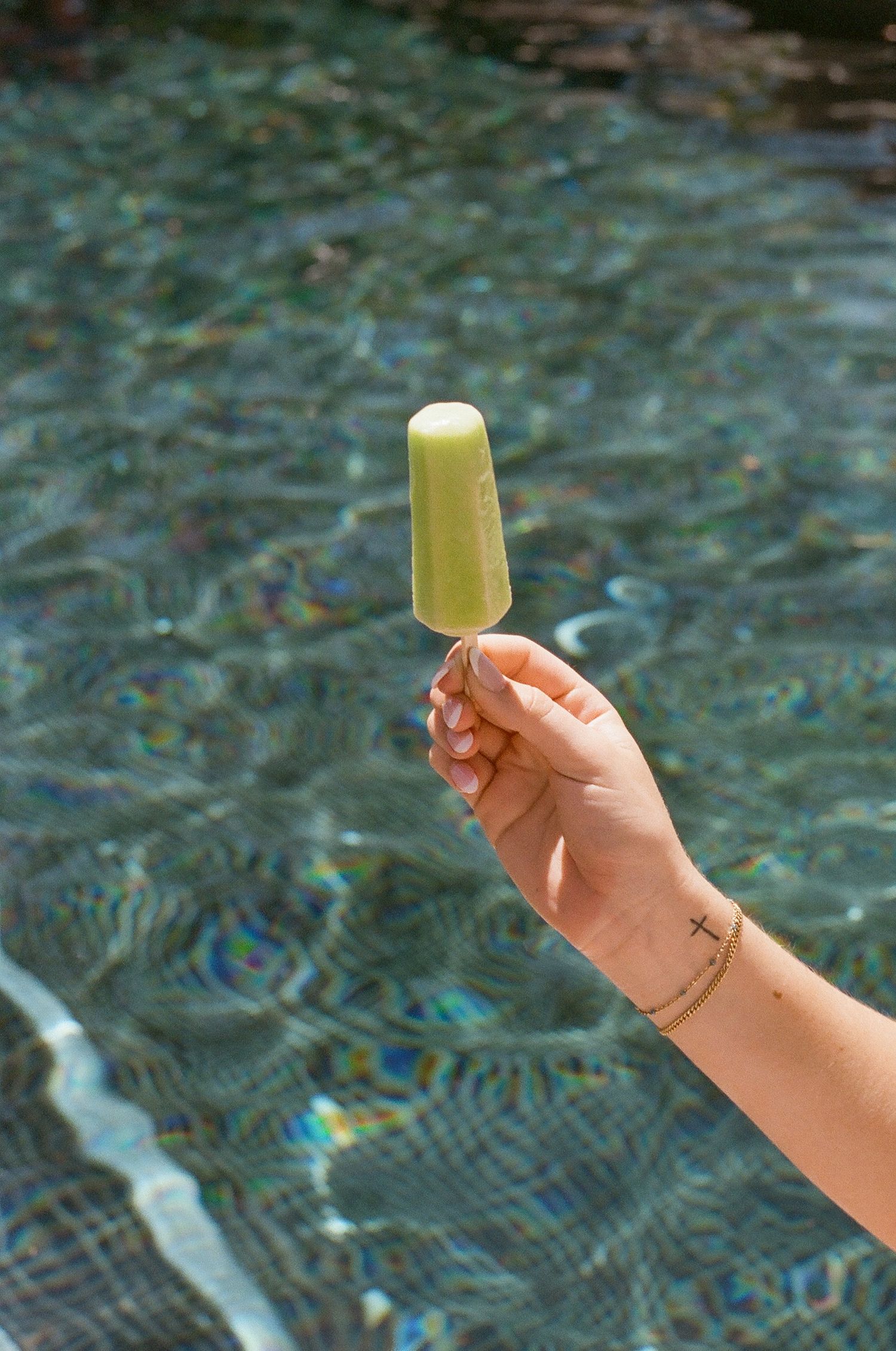 A hand holds up a yellow popsicle against a shimmering blue pool water background.