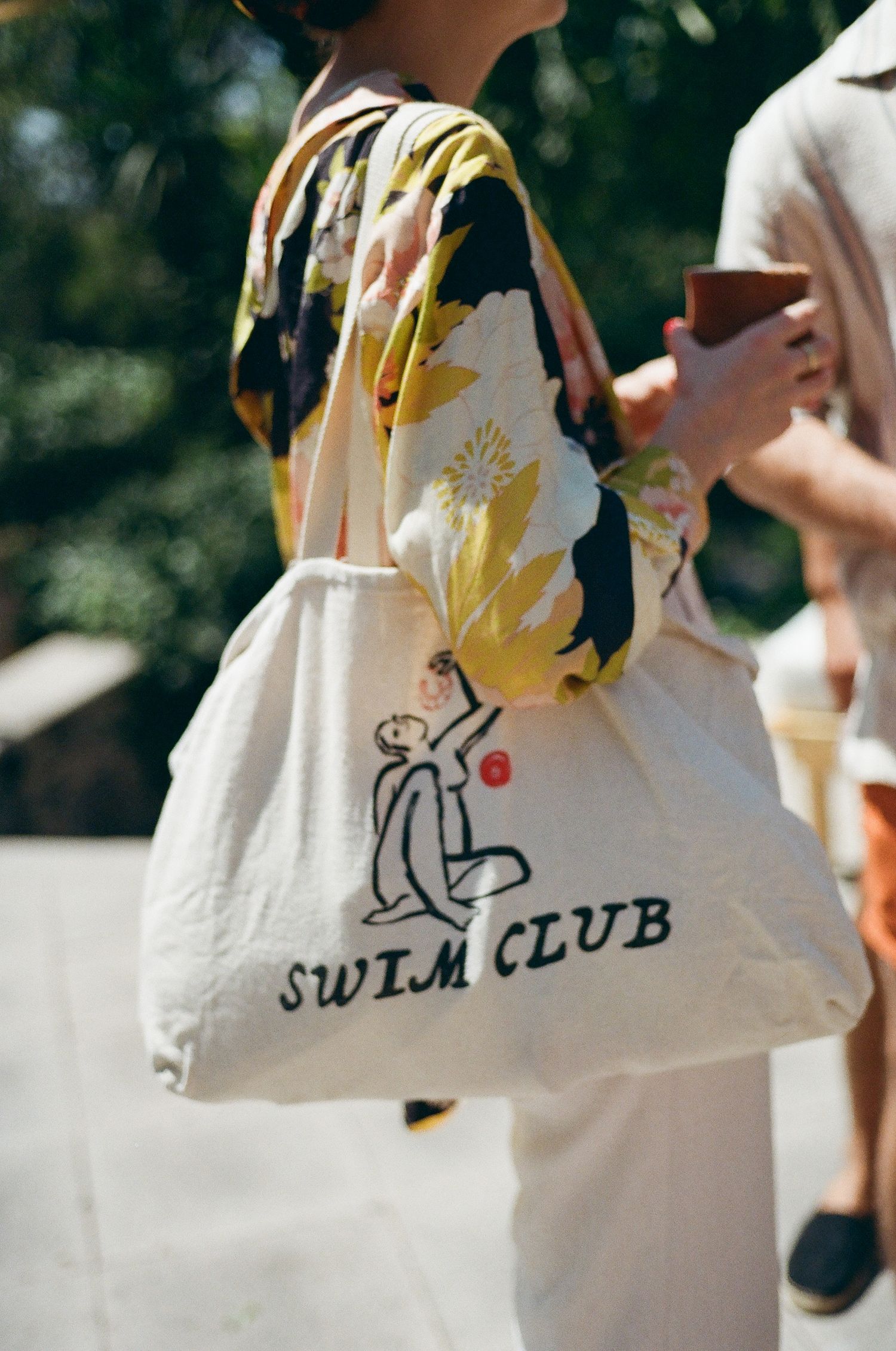 Row of white tote bags with swim club text and designs displayed outdoors.
