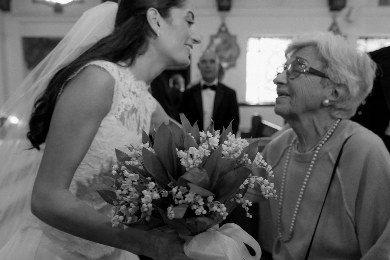 Bride in lace gown smiling at elderly woman holding bouquet of lily of the valley at wedding ceremony.
