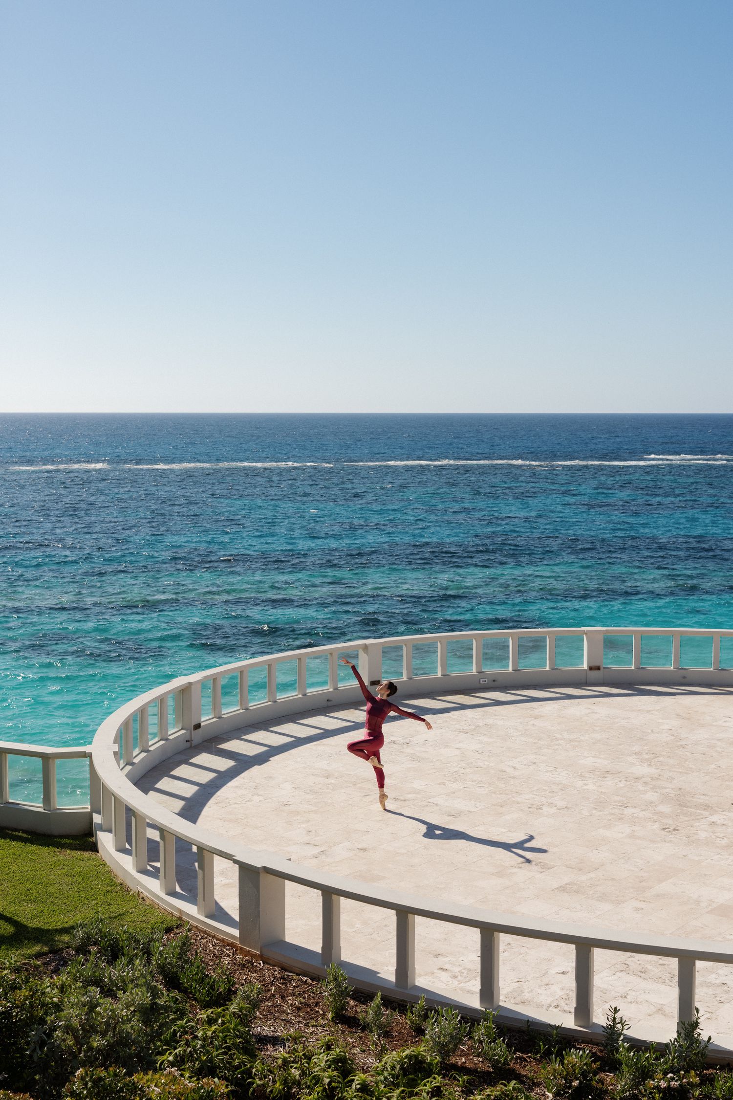 The image depicts a person standing on a circular platform overlooking a body of turquoise water against a clear blue sky.