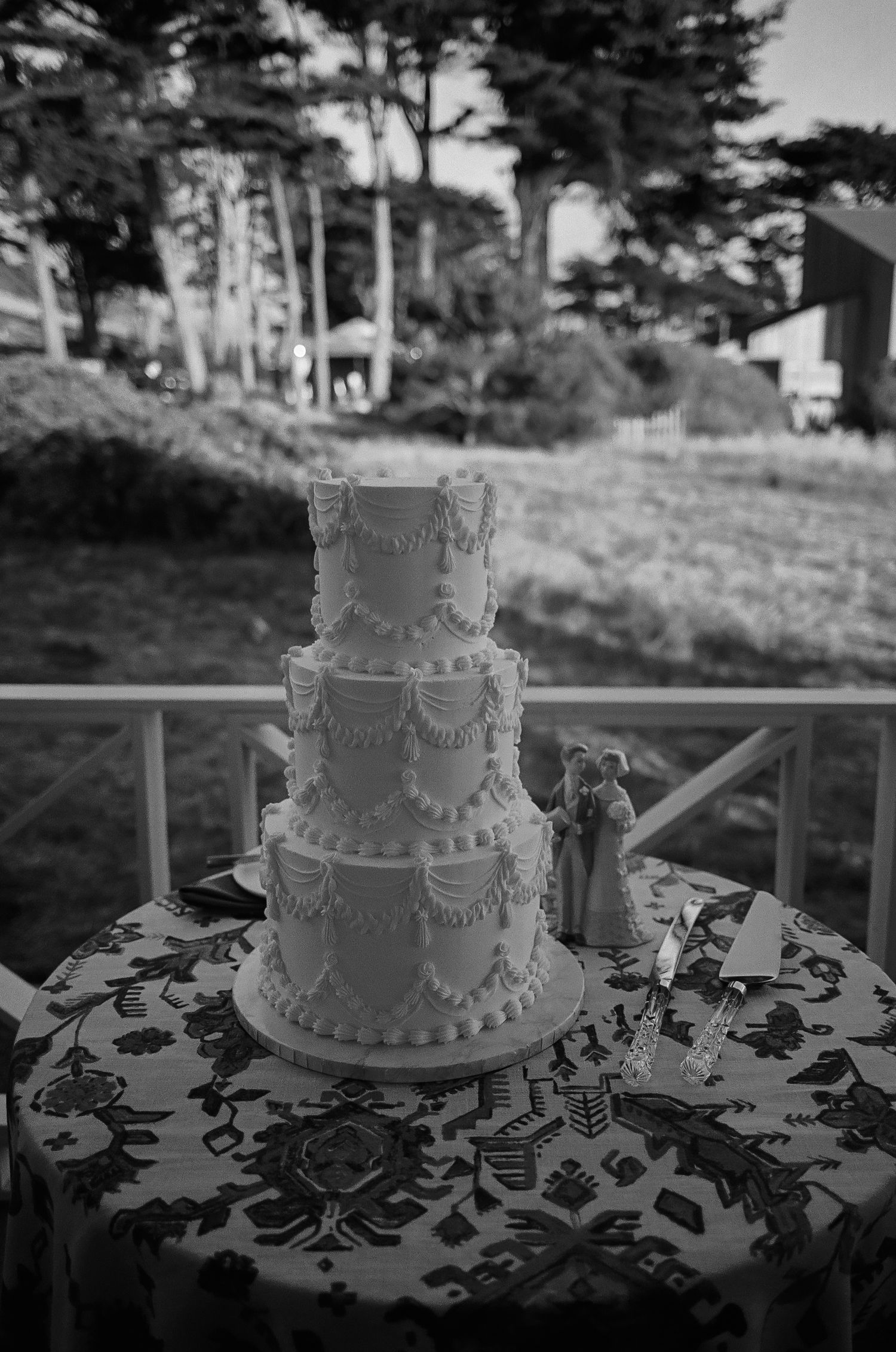 Elegant white tiered wedding cake displayed on a patterned tablecloth outdoors.