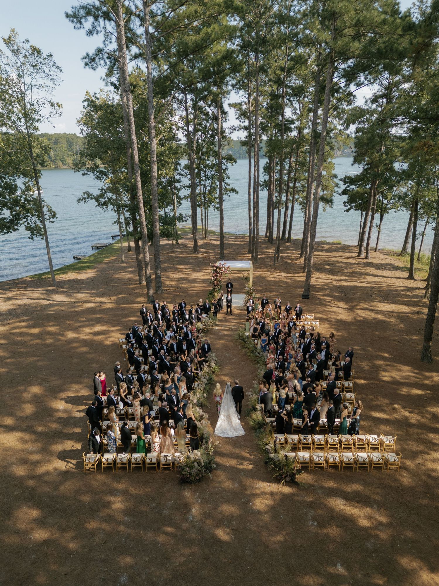 Wedding ceremony setup with guests arranged in symmetrical rows among lakeside pine trees.