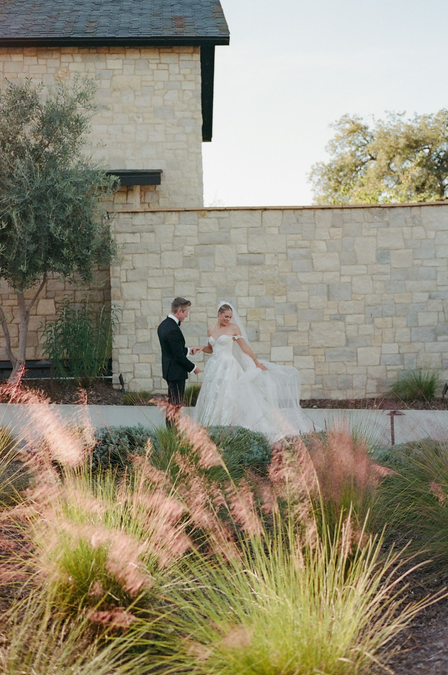 A couple stands together on stone path surrounded by ornamental grasses near a stone building.