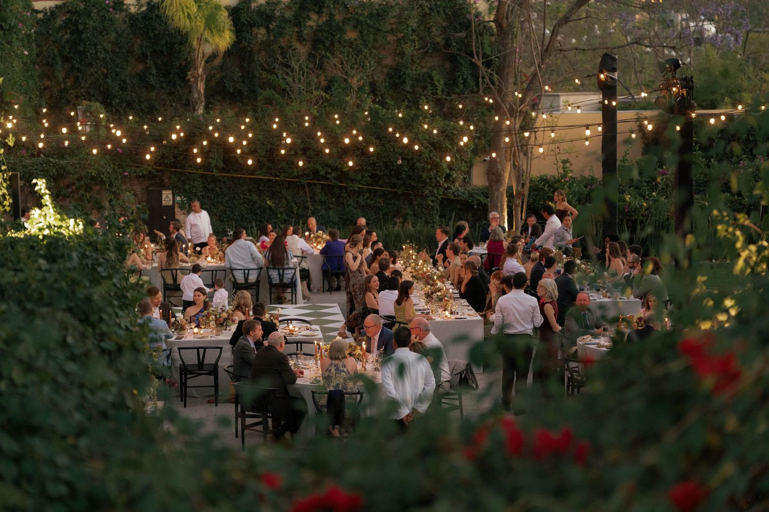 Outdoor wedding reception at dusk with string lights creating a warm ambiance in a garden setting.