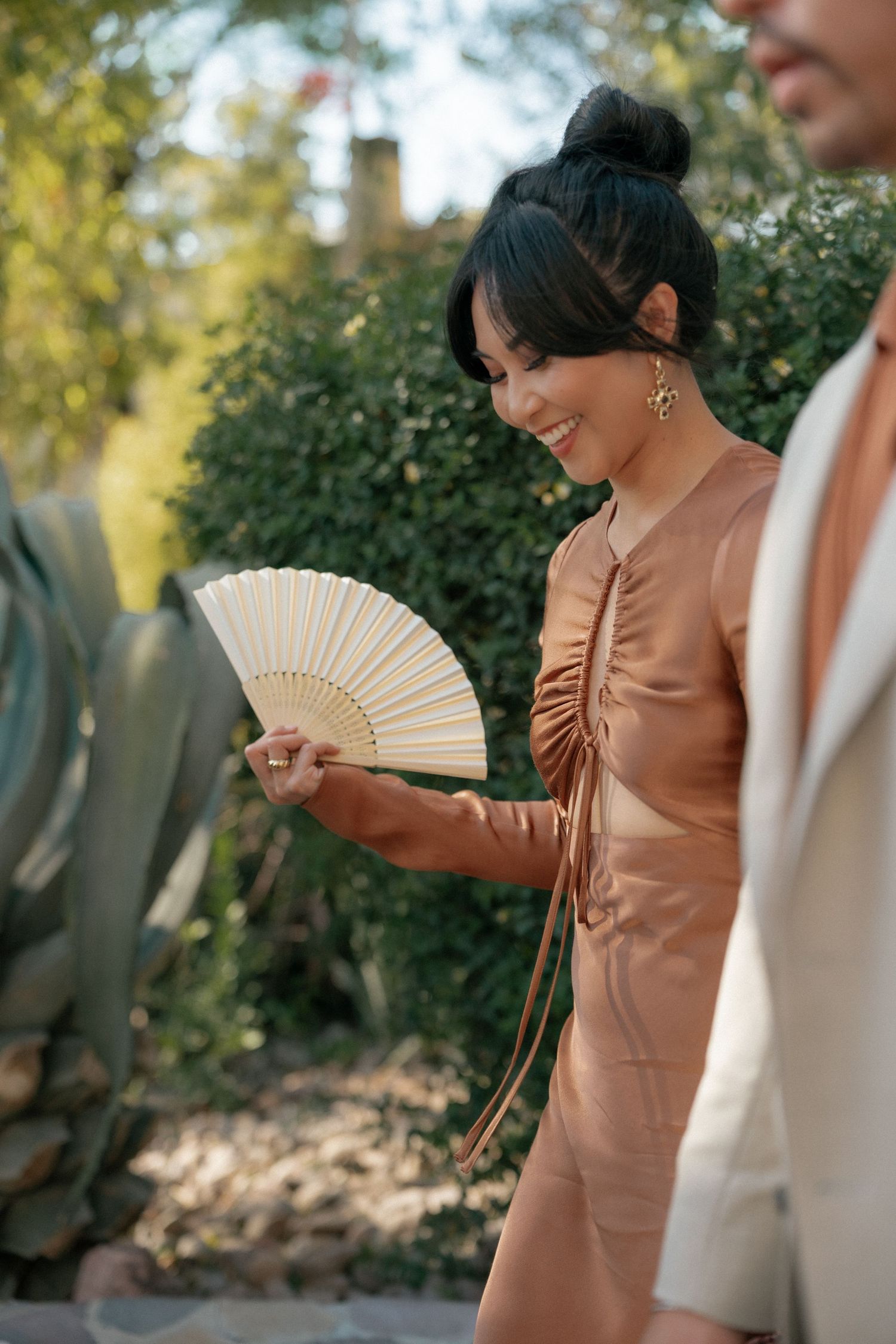Wedding guest in copper-colored dress holds decorative fan at outdoor reception.