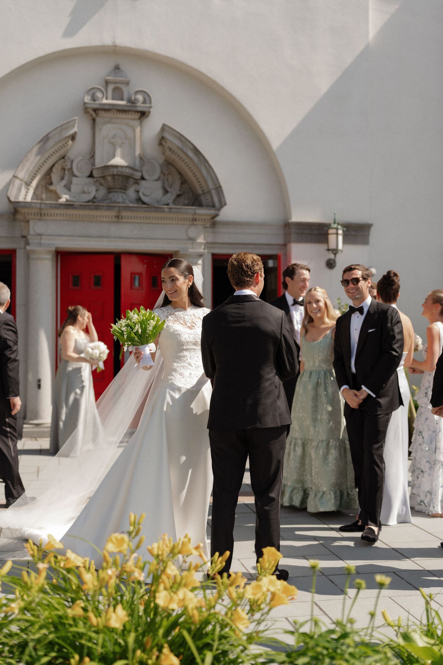 Bride in white gown holds bouquet outside church with red doors, greeted by wedding guests in formal attire.