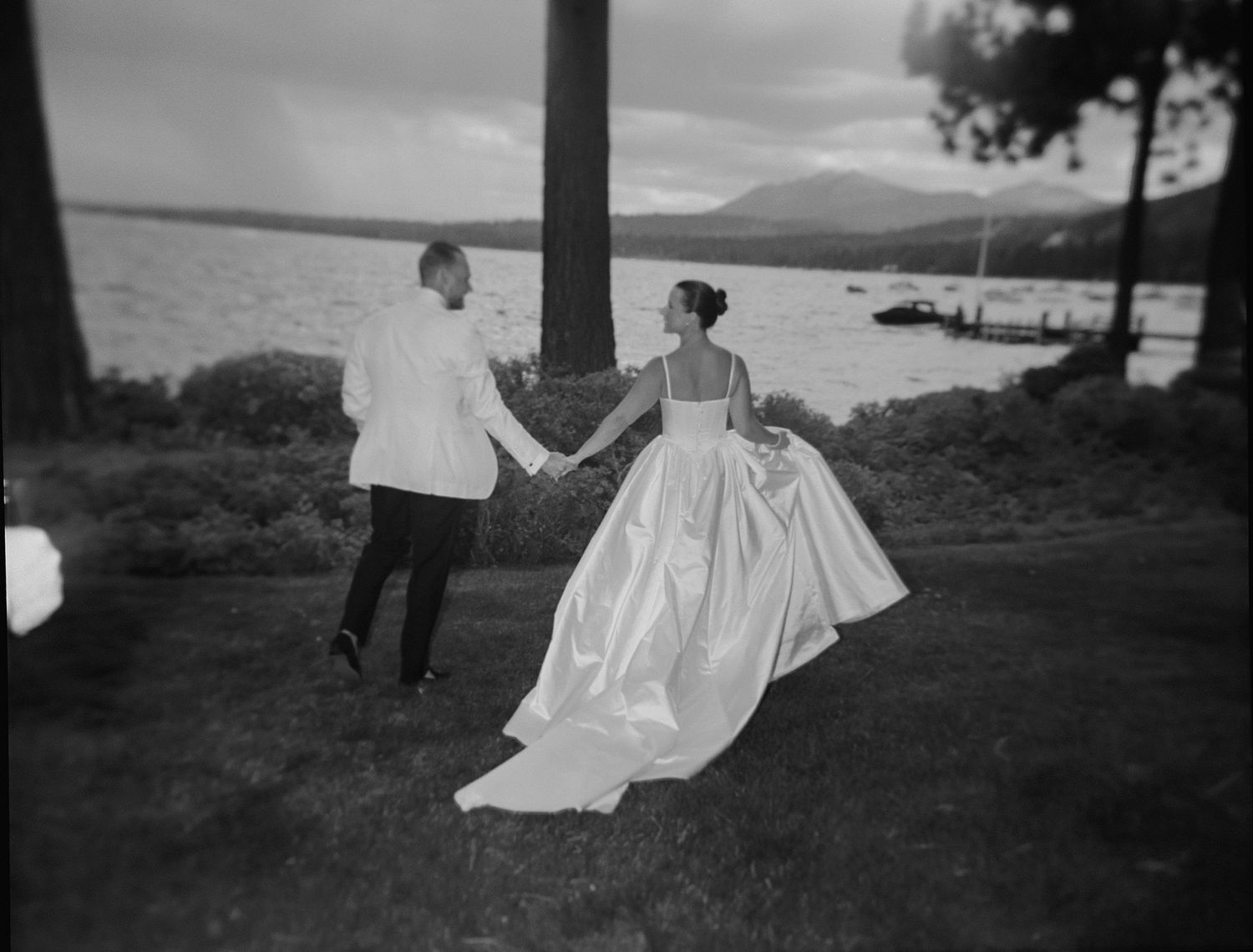 A couple walks hand in hand across a scenic lakeside lawn in formal attire.