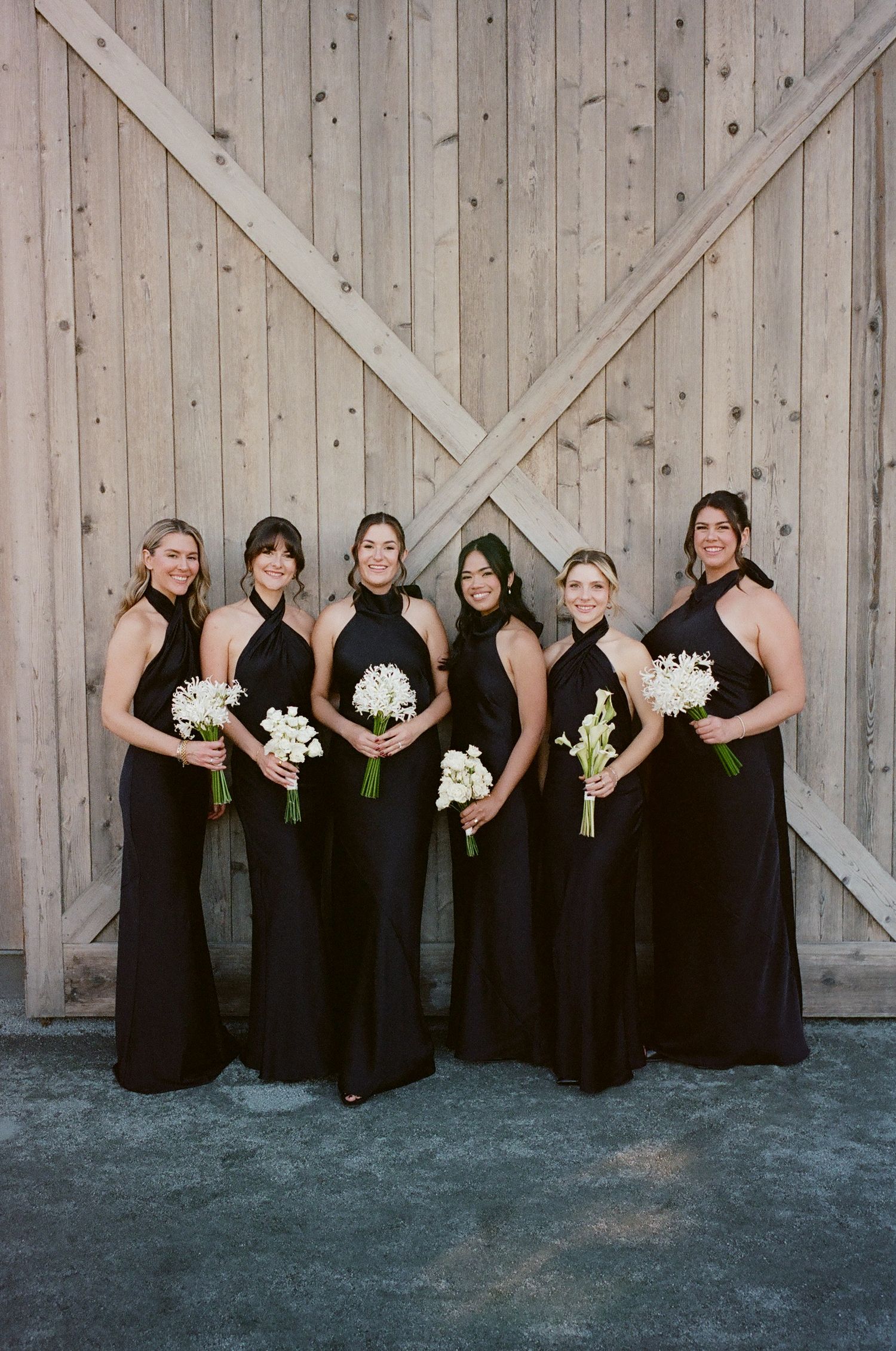 Wedding party members in black dresses pose with white bouquets against a wooden barn backdrop.