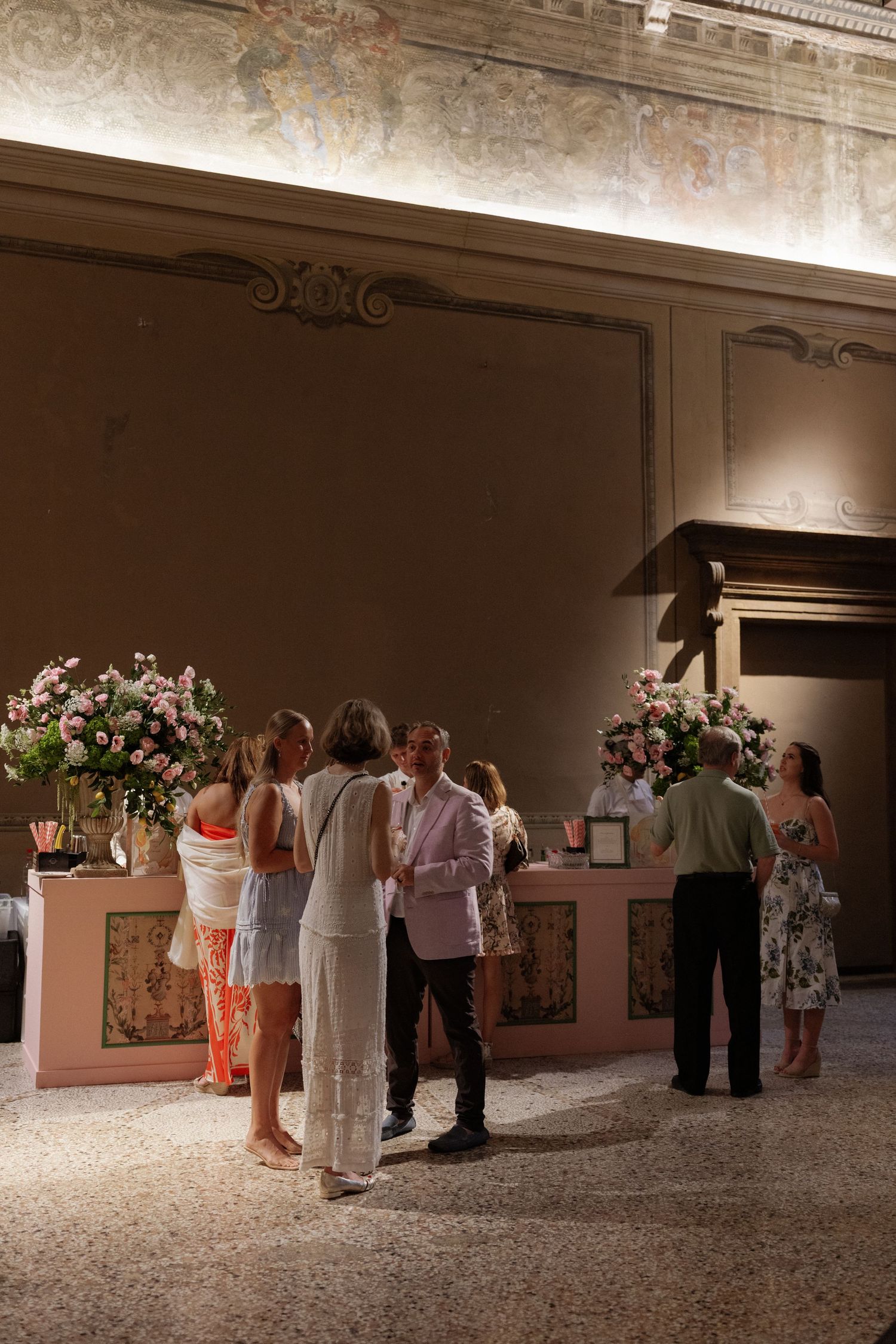 Elegant indoor reception with guests mingling near floral arrangements in a ornate room with decorative murals.